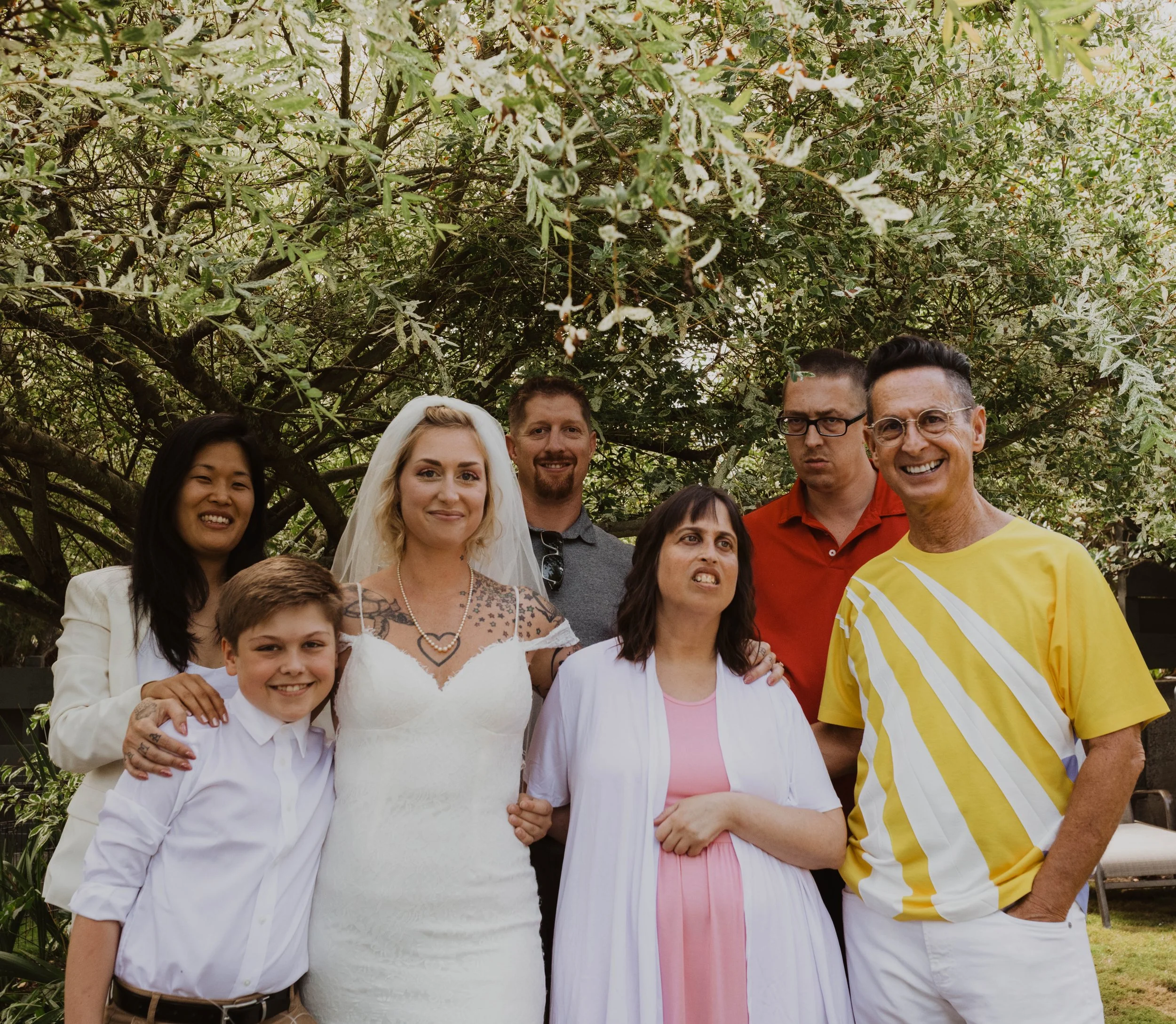 A group of seven people, including a bride in a white wedding dress with a veil, and six others, standing outdoors under a leafy tree, smiling and posing for a photo. Seattle, WA wedding photography.