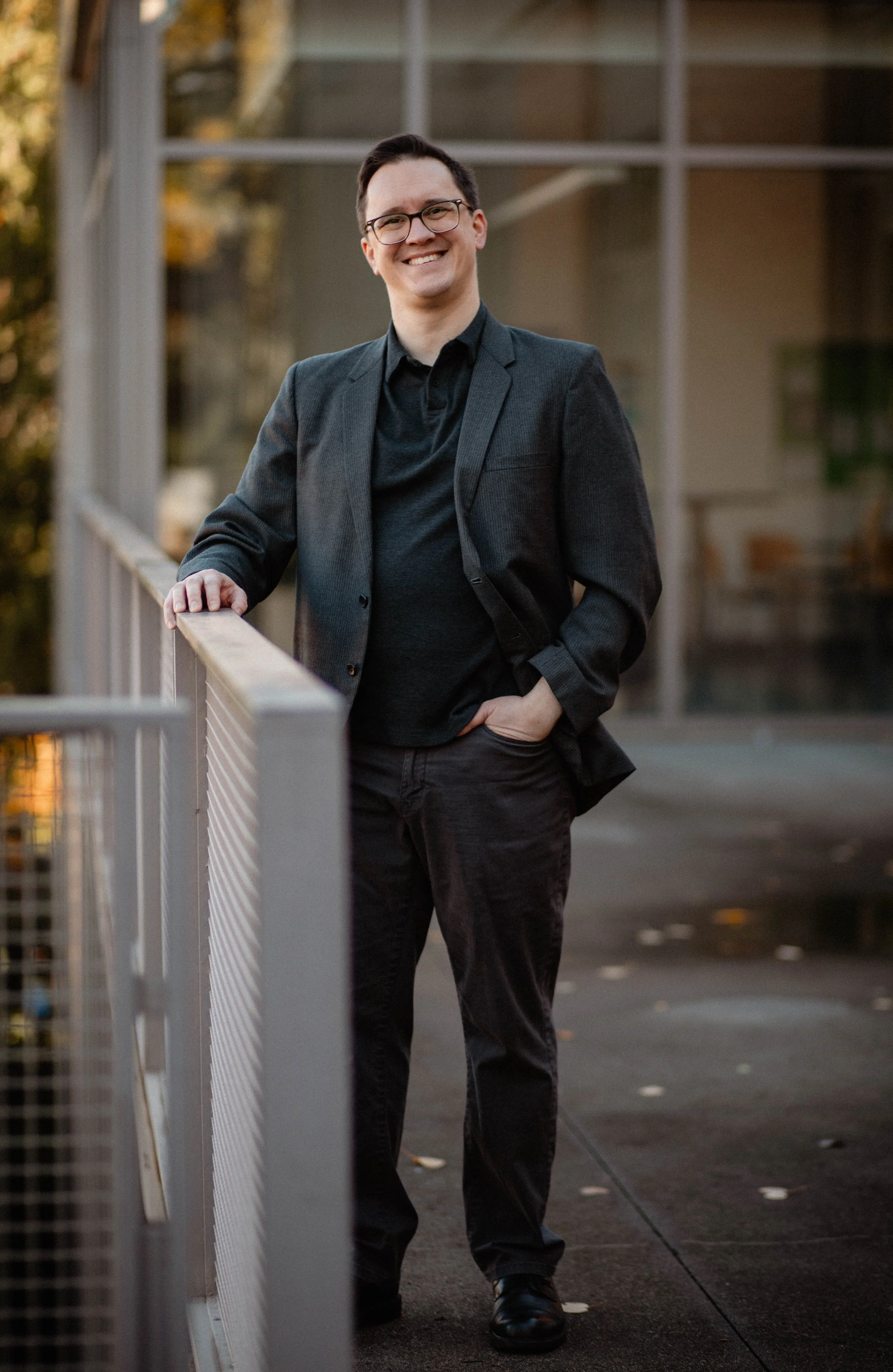 A man in a dark blazer and glasses smiling outdoors on a balcony railing. Seattle professional head shot photography