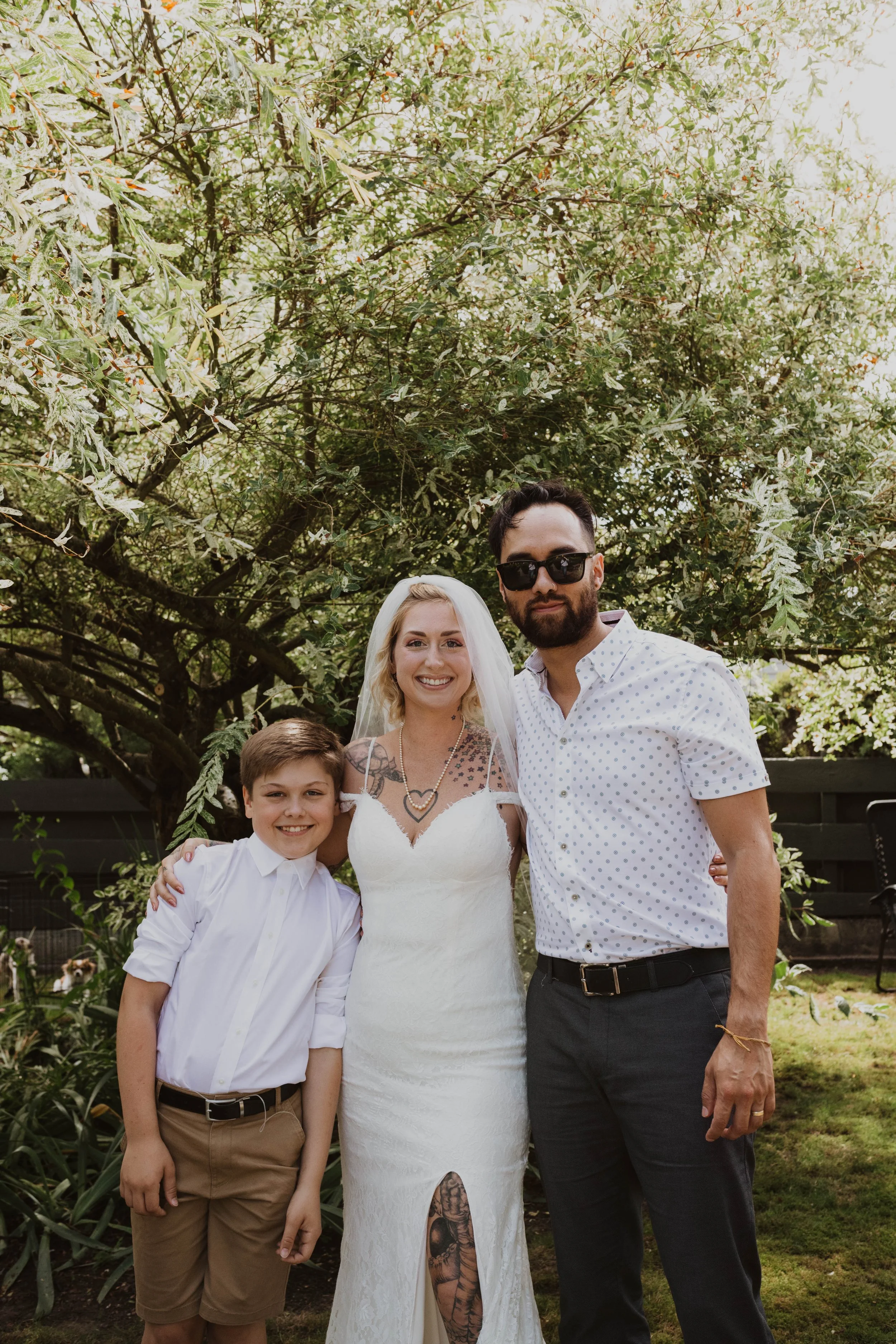 A bride and two males, possibly family, standing outdoors under a large tree with green foliage, smiling for a photo on a sunny day. Seattle, WA wedding photography.