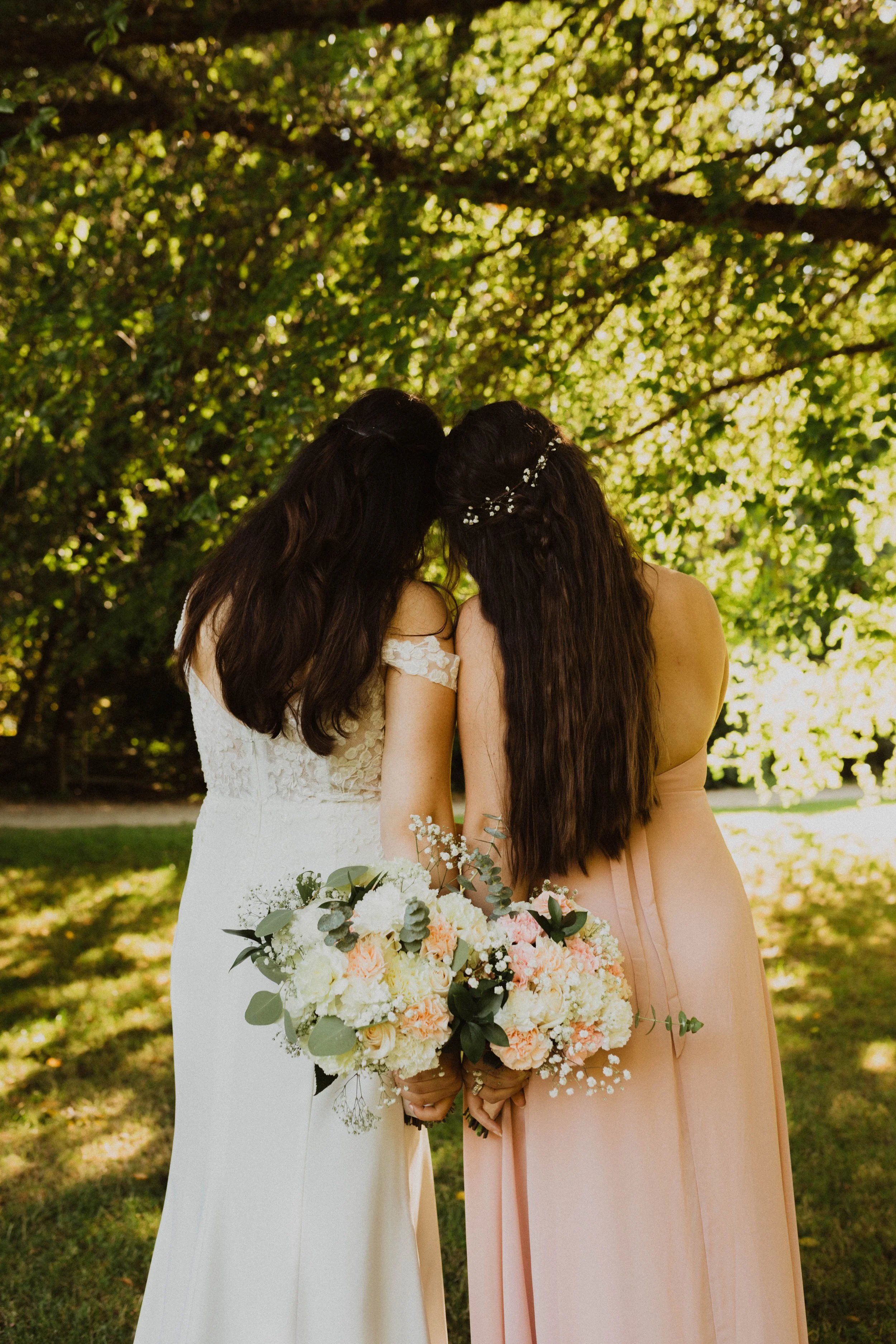 Two women in wedding attire standing outdoors under a tree, holding bouquets of flowers, with their heads touching. Seattle, WA wedding photography.
