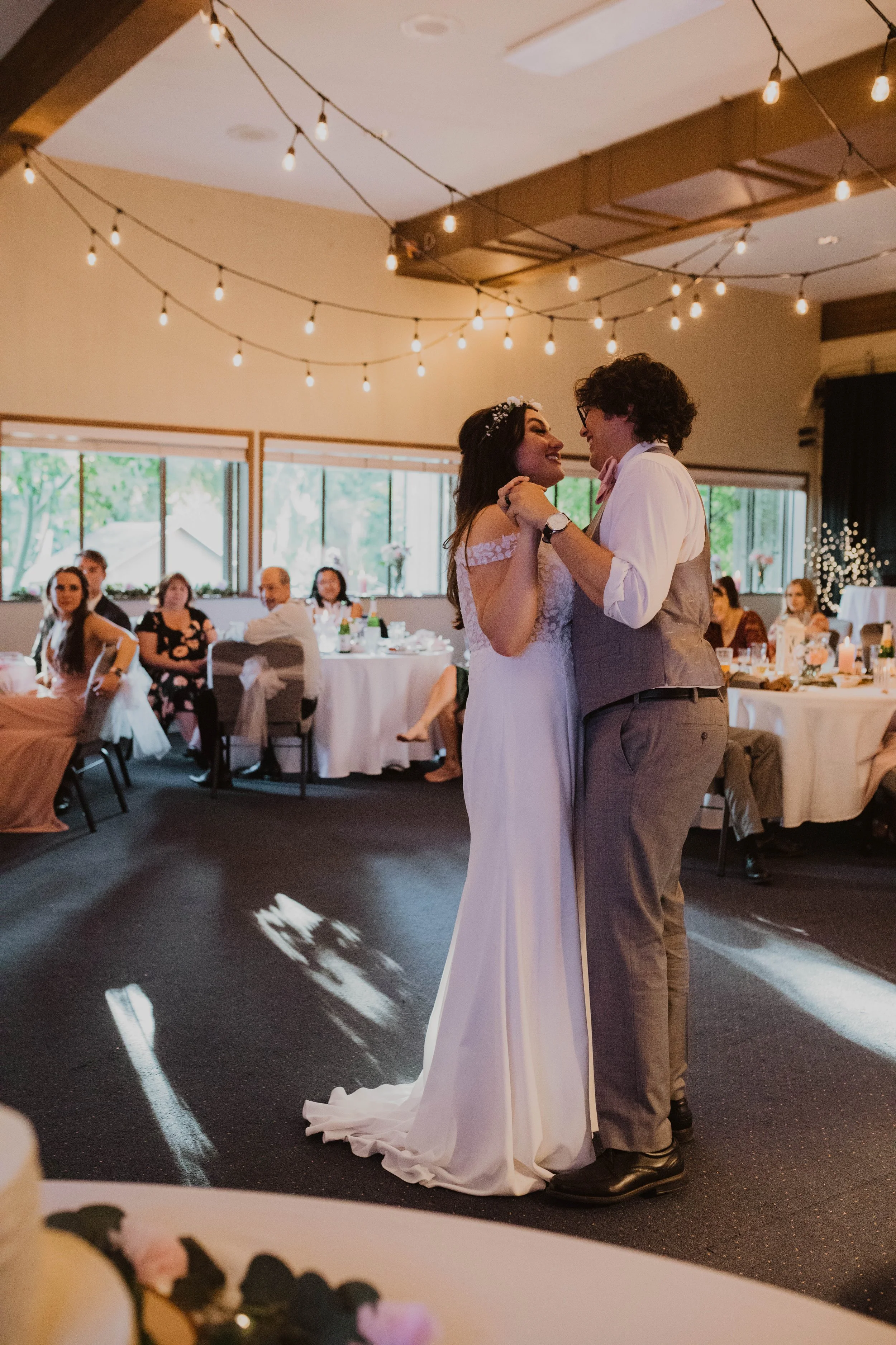 A bride and groom dance together at their wedding reception, with guests seated at tables watching. The room is decorated with string lights and large windows. Seattle, WA wedding photography.