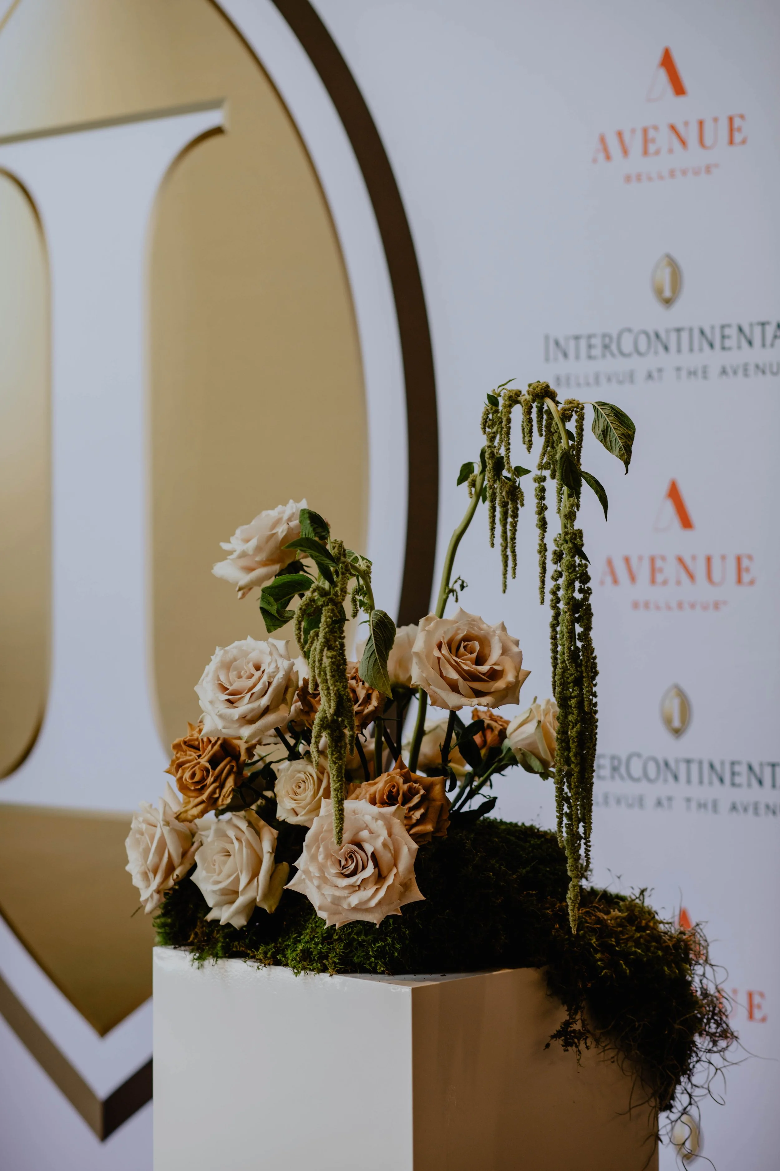 A floral arrangement with cream and brown roses on a white pedestal in front of a backdrop with logos and text for Avenue Bellevue and InterContinental Bellevue at The Avenue. Seattle event photography