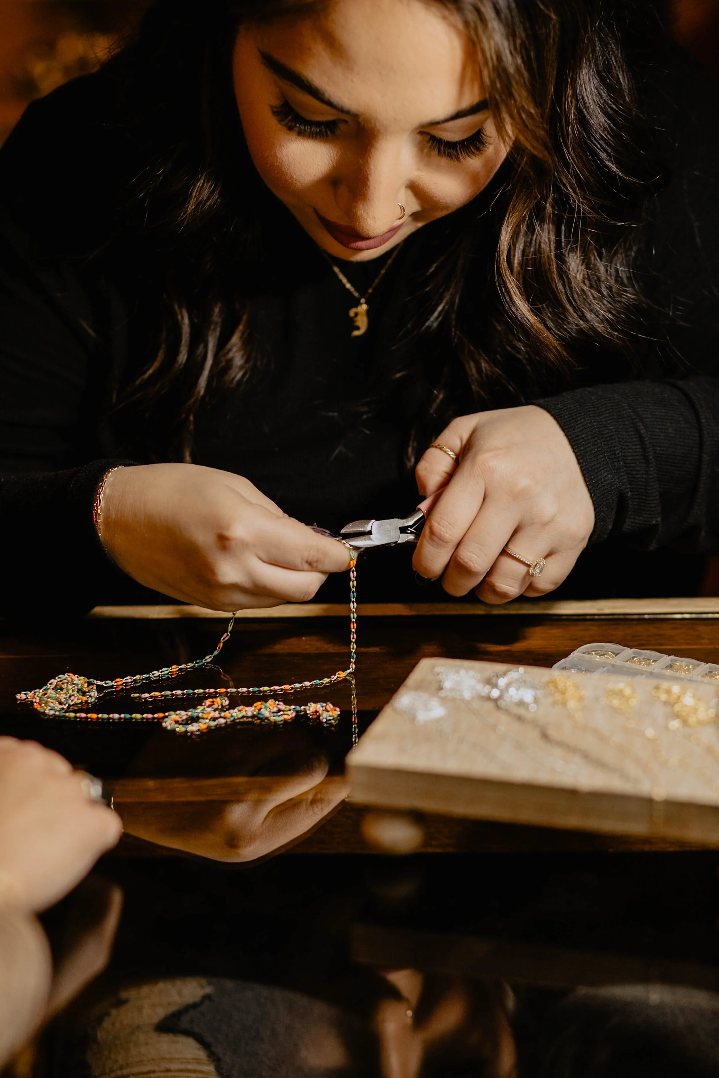 A woman with dark wavy hair and jewelry working on a colorful beaded necklace with pliers at her workspace, with a box of jewelry on the table. Seattle professional head shot photography
