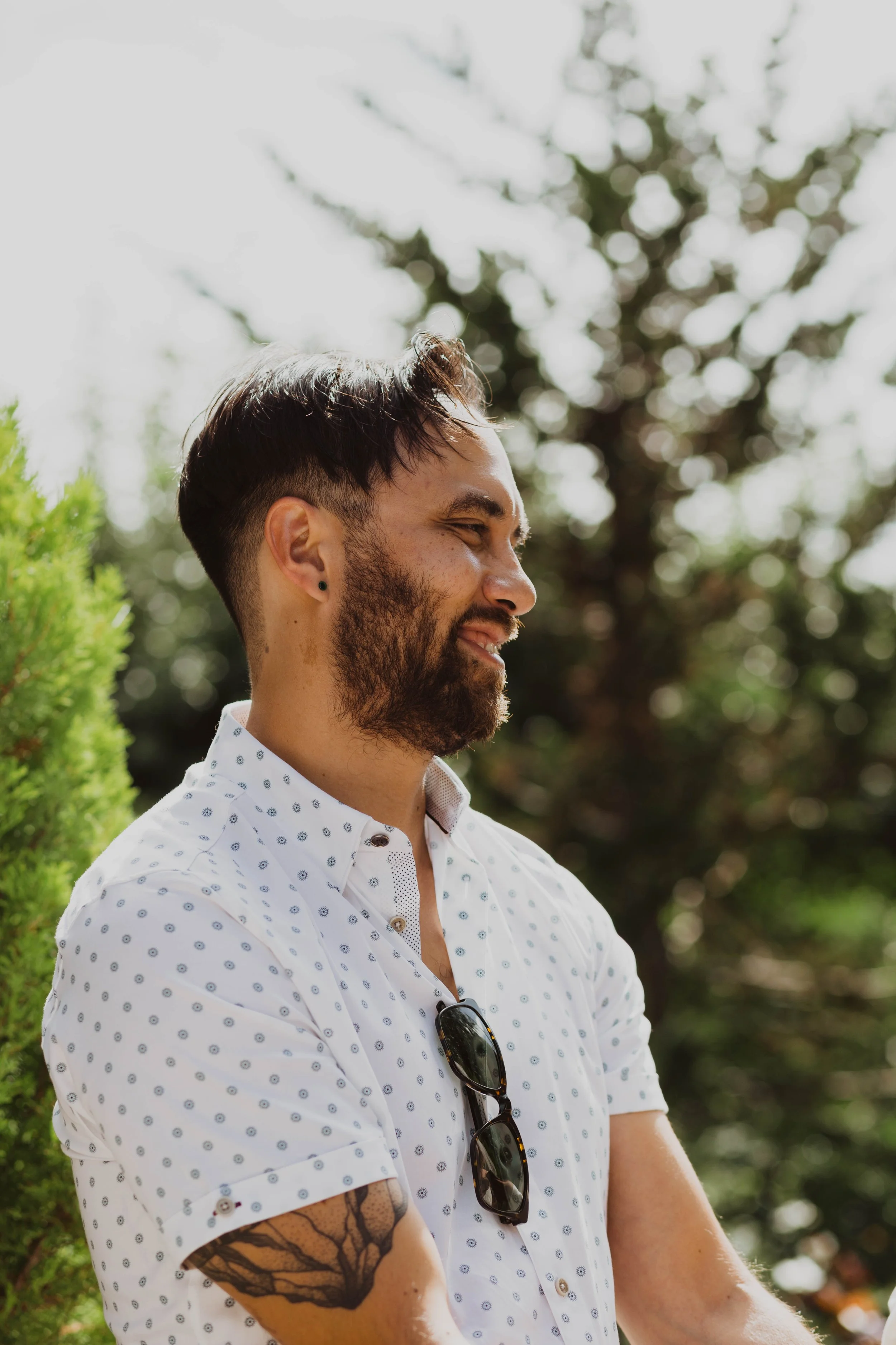 A man with dark hair and a beard smiling outdoors, wearing a white shirt with small patterns, sunglasses hanging on his shirt, and a tattoo on his arm, with trees and sunlight in the background. Seattle, WA wedding photography.