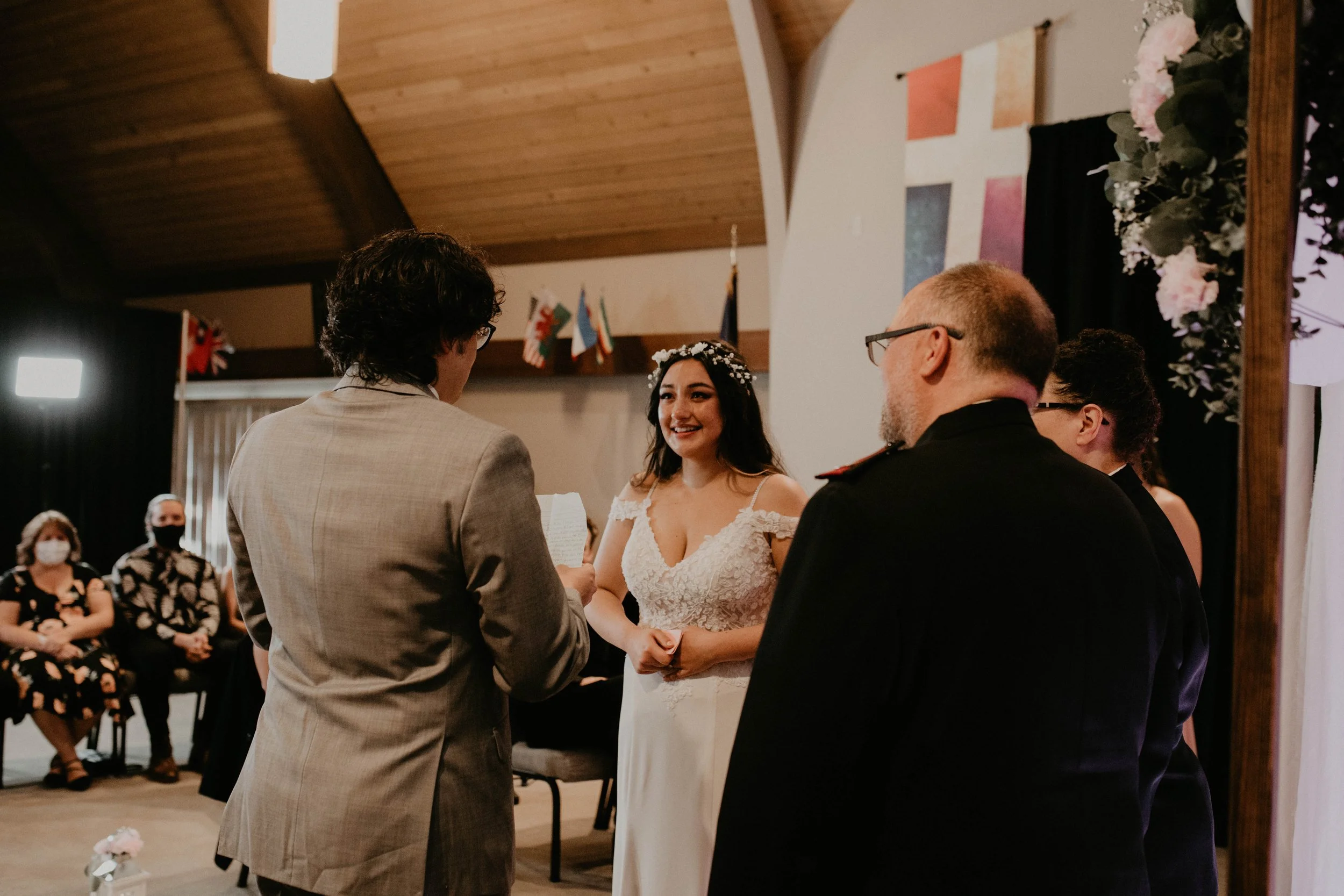 A wedding ceremony with a bride in a white lace dress smiling as she looks at the groom, who is reading vows. The officiant is in front of them, and guests are seated in the background wearing masks. Seattle, WA wedding photography.
