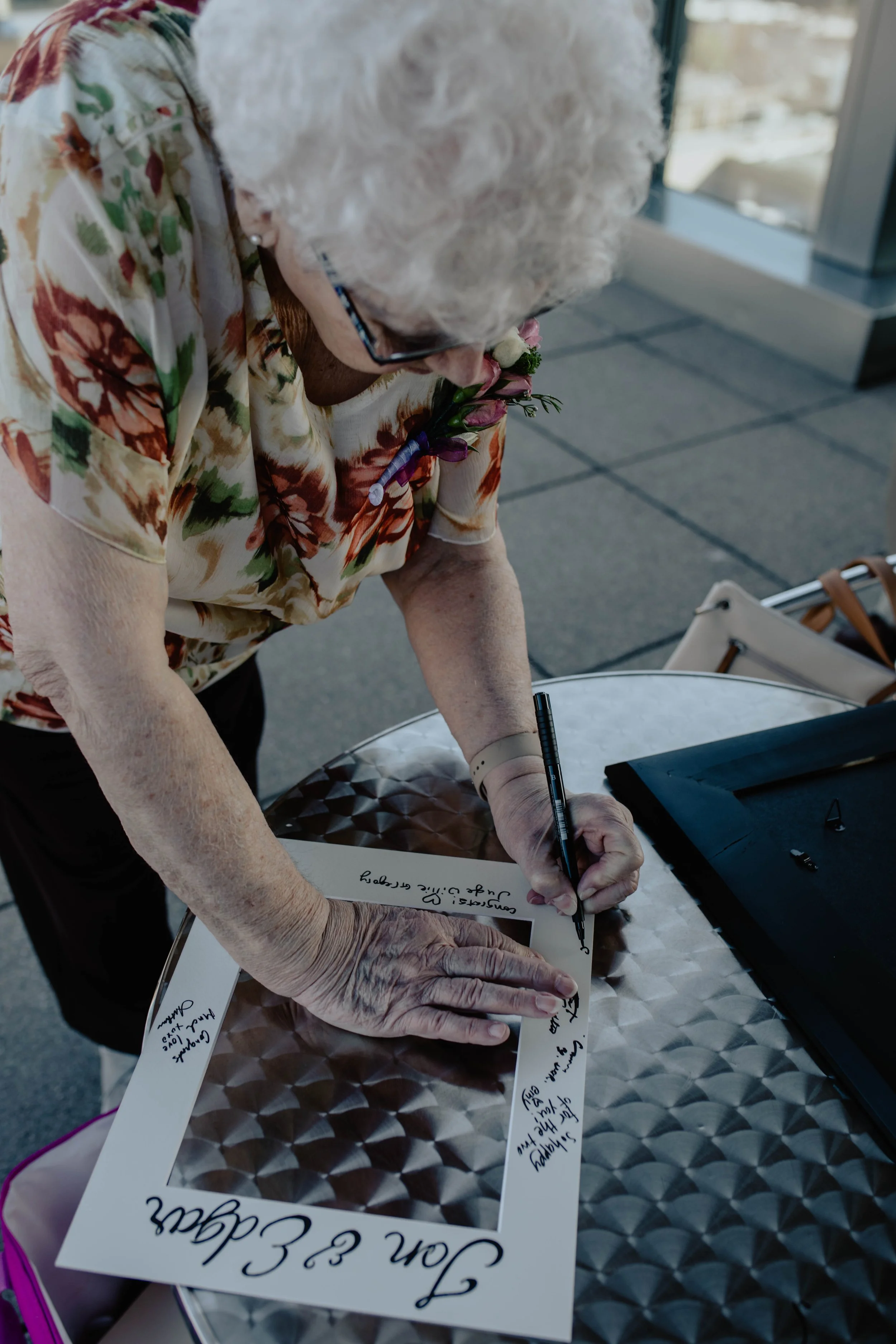 An elderly woman with white curly hair, wearing glasses and a floral blouse, is signing a framed photo or certificate. She appears to be at an outdoor event or celebration, standing at a metallic table with a black tablet or book nearby. Seattle Muni