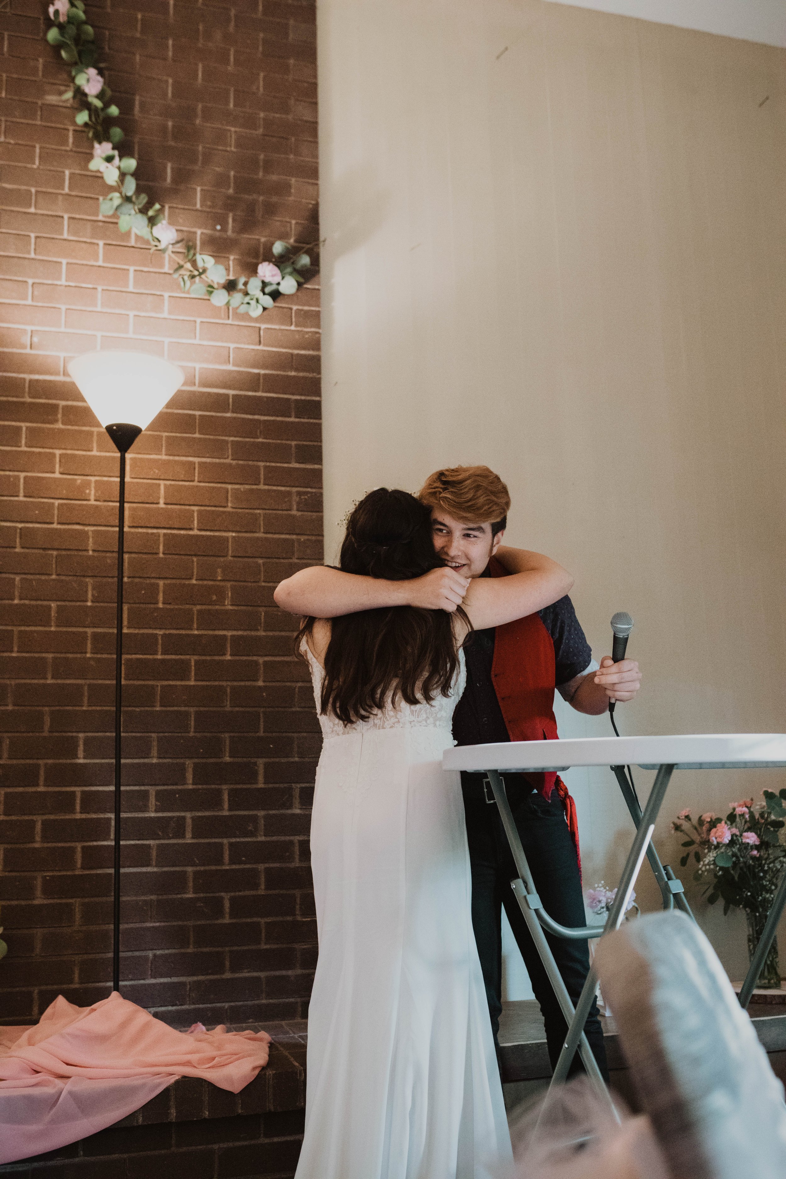A young woman in a white dress hugging a young man with a microphone at a celebration or gathering indoors, with a brick wall, a standing lamp, and floral decorations in the background. Seattle, WA wedding photography.