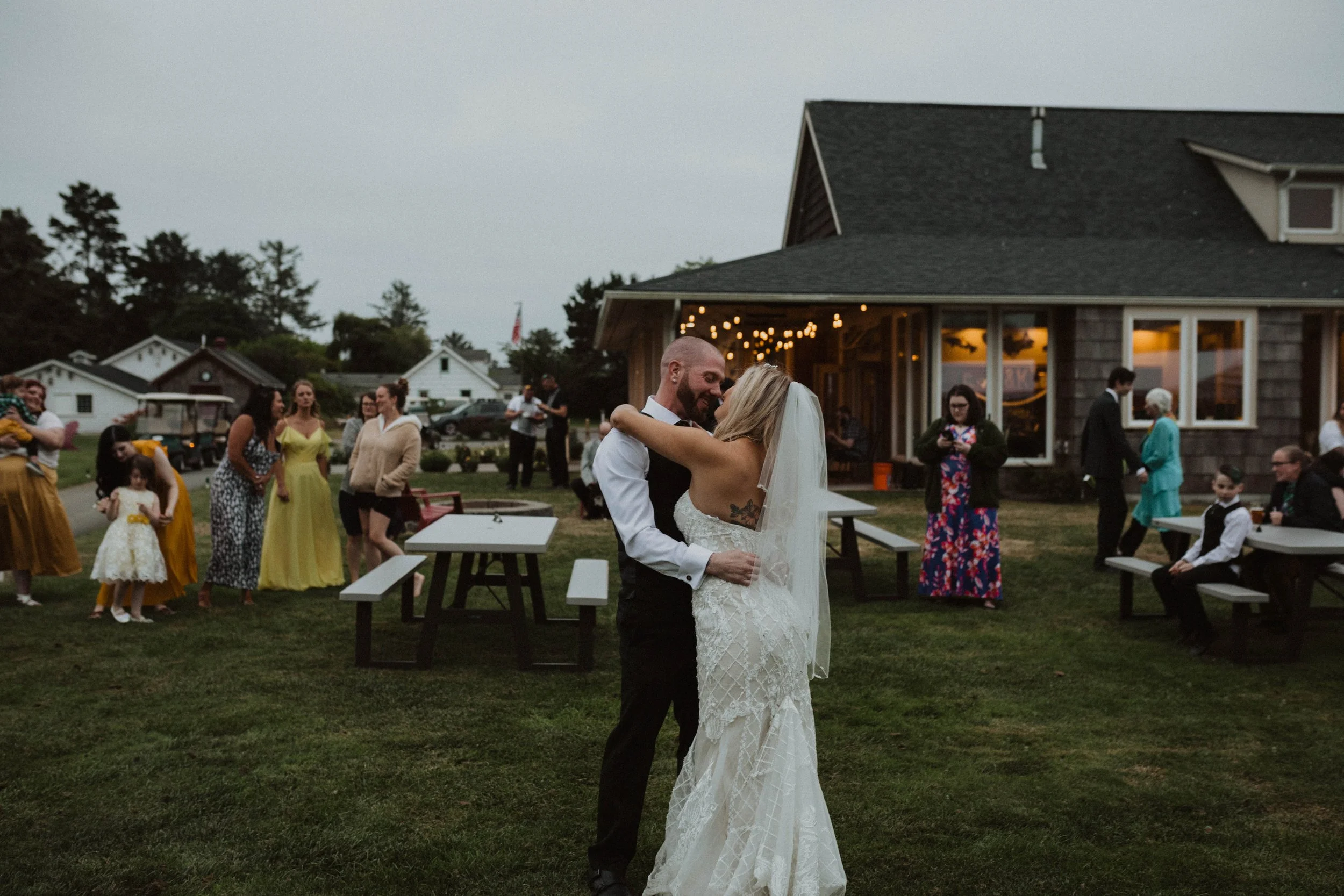 A bride and groom dancing outdoors at their wedding reception, surrounded by guests, with a rustic building and string lights in the background during early evening. Long Beach, WA wedding photography.