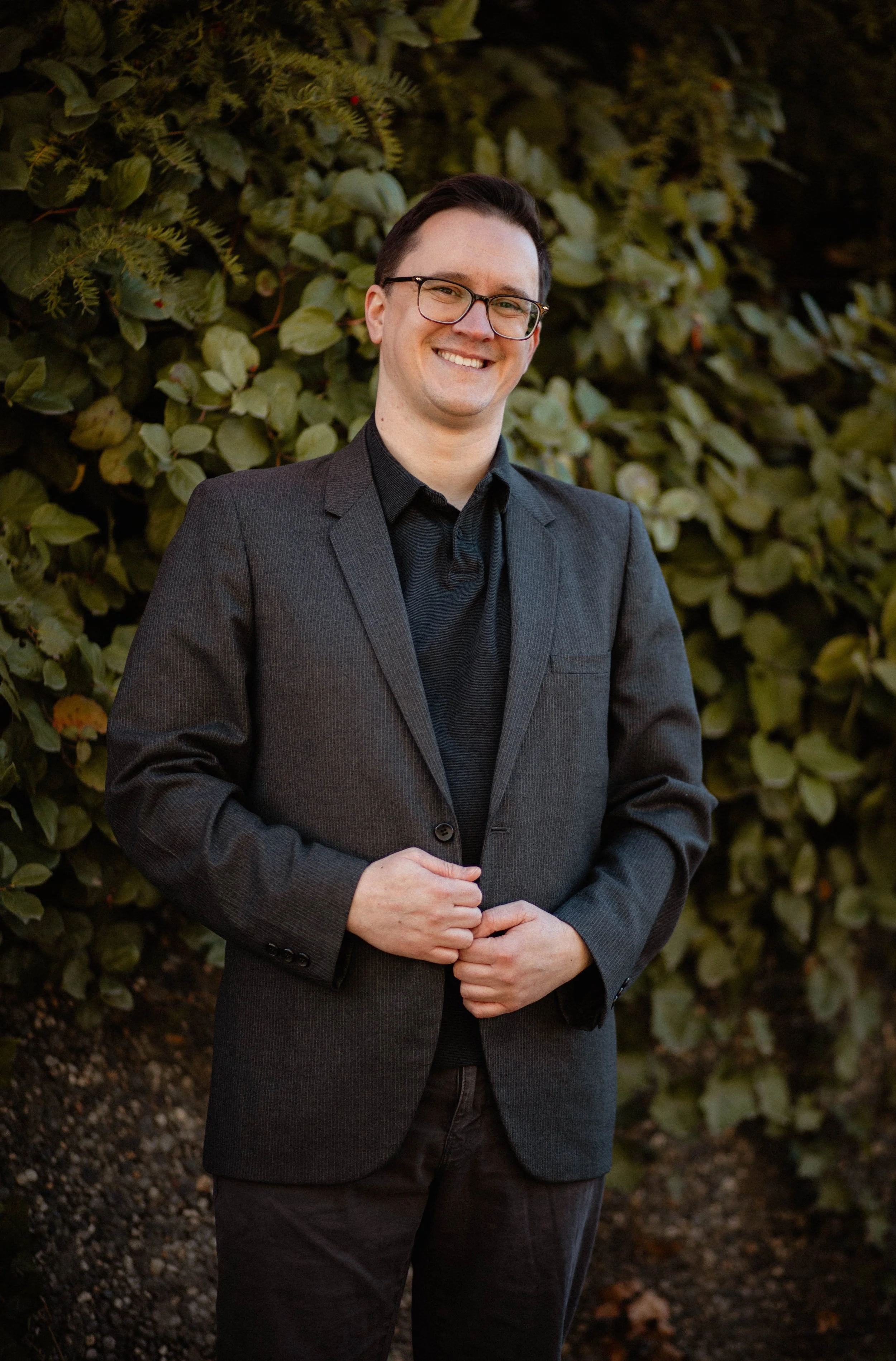 A man in glasses wearing a dark blazer and shirt, smiling and standing outdoors in front of green leafy bushes. Seattle professional head shot photography