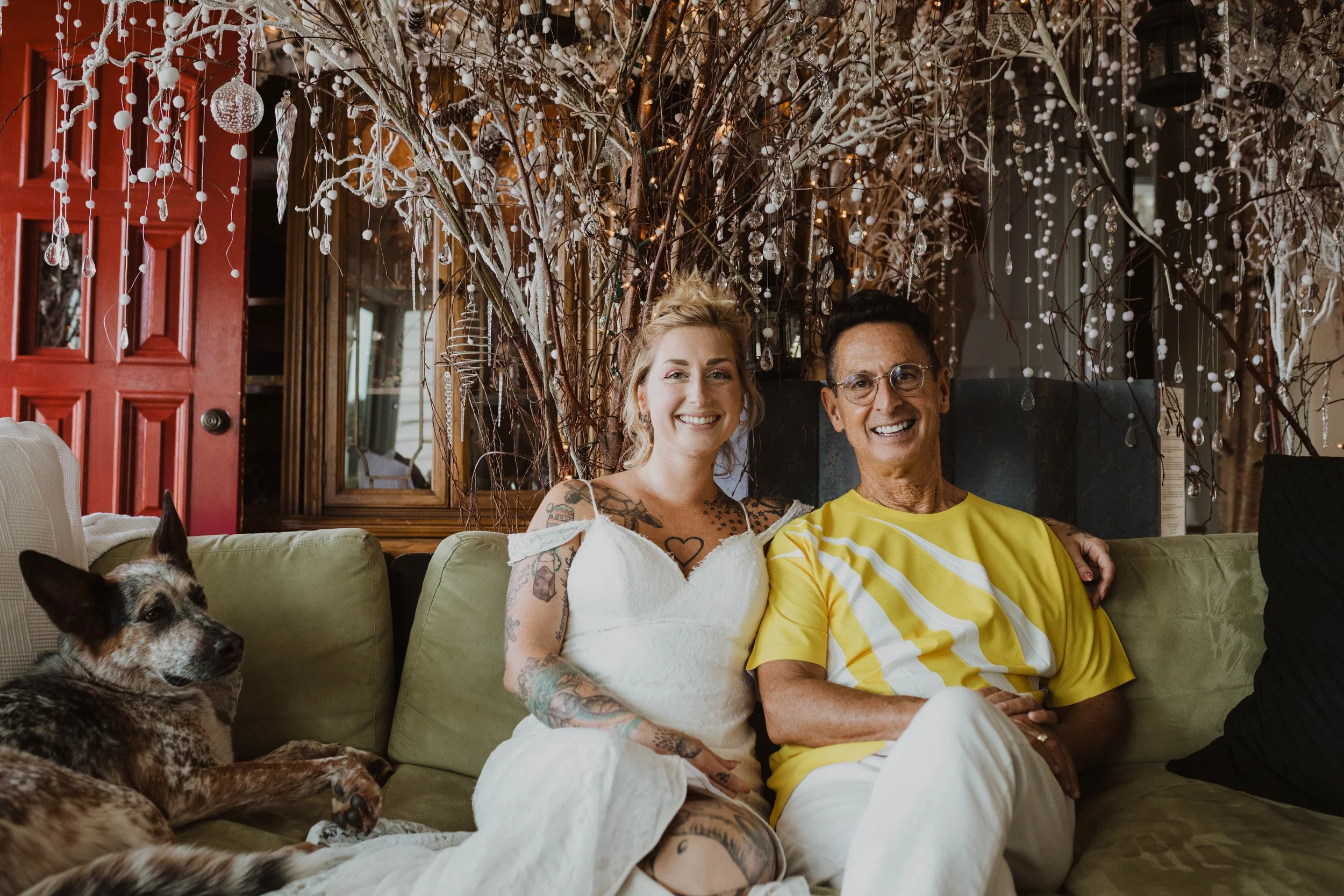 A woman with tattoos and blond hair, dressed in white, and a man with glasses and a yellow shirt, sitting on a green couch, smiling, with a dog lying beside them. They are in a room decorated with branches, white ornaments, and fairy lights. Seattle,