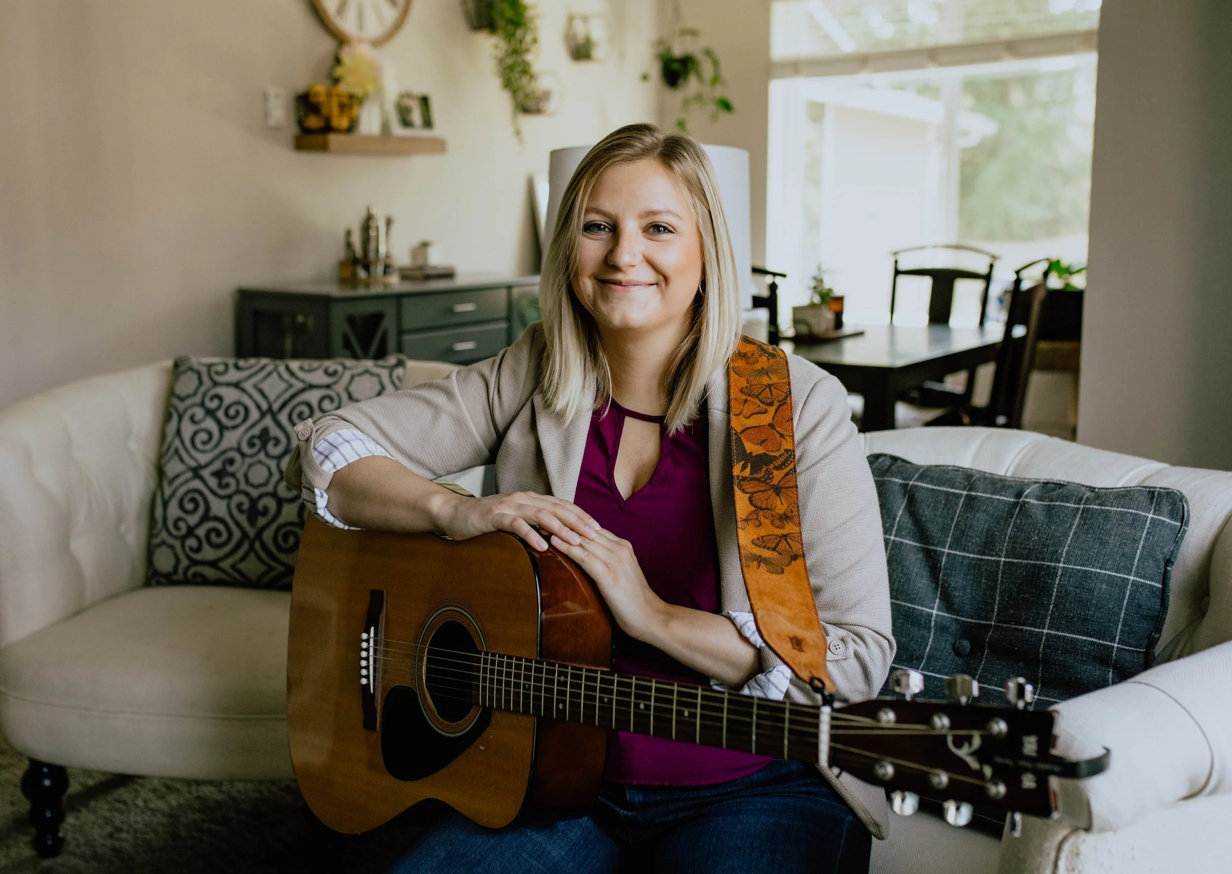 A woman sitting on a white sofa holding an acoustic guitar, smiling at the camera in a cozy living room with plants and decorative items. Seattle professional head shot photography