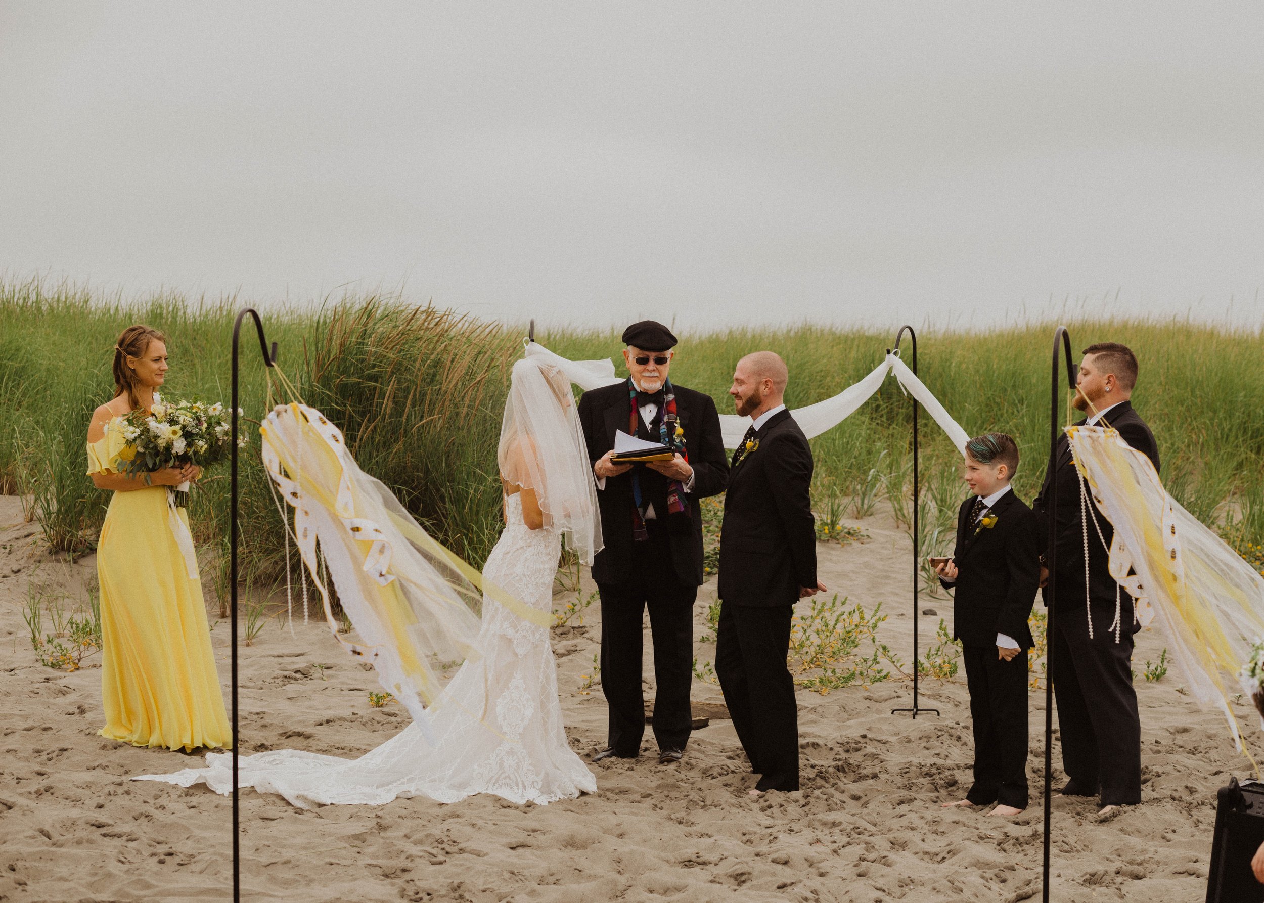 A beach wedding ceremony with a bride and groom standing before an officiant, surrounded by flower girl and ring bearers, with decorative fabric and ribbons in a sand and grass setting under an overcast sky. Long Beach, WA wedding photography.