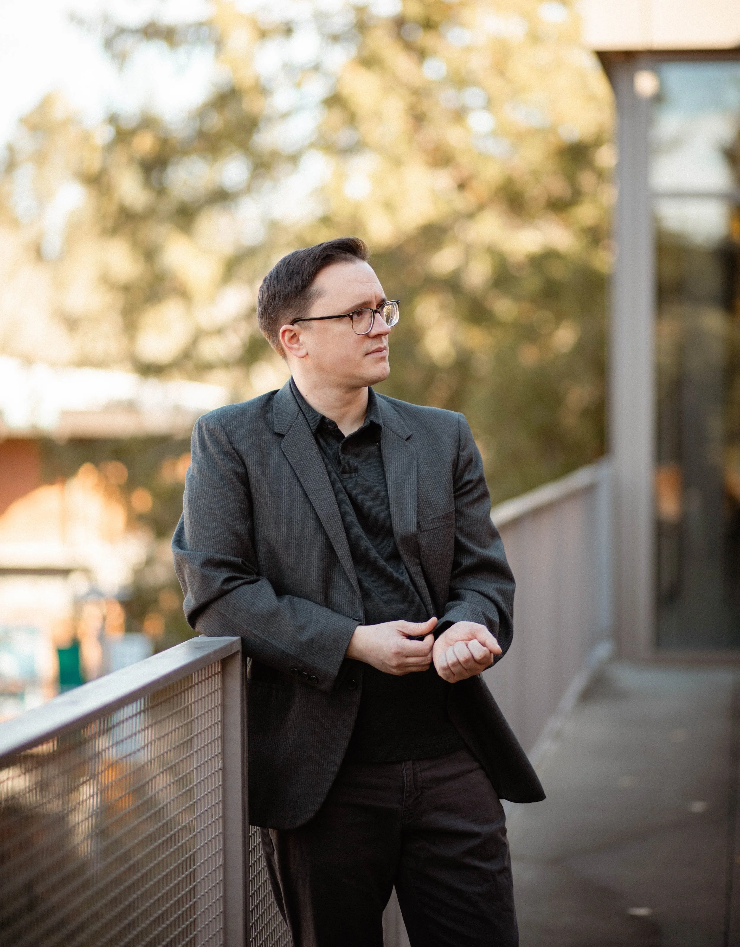A man wearing glasses and a dark gray blazer leans against a metal railing outdoors during daytime. He is looking to the side with a contemplative expression, surrounded by blurred trees and buildings. Seattle professional head shot photography