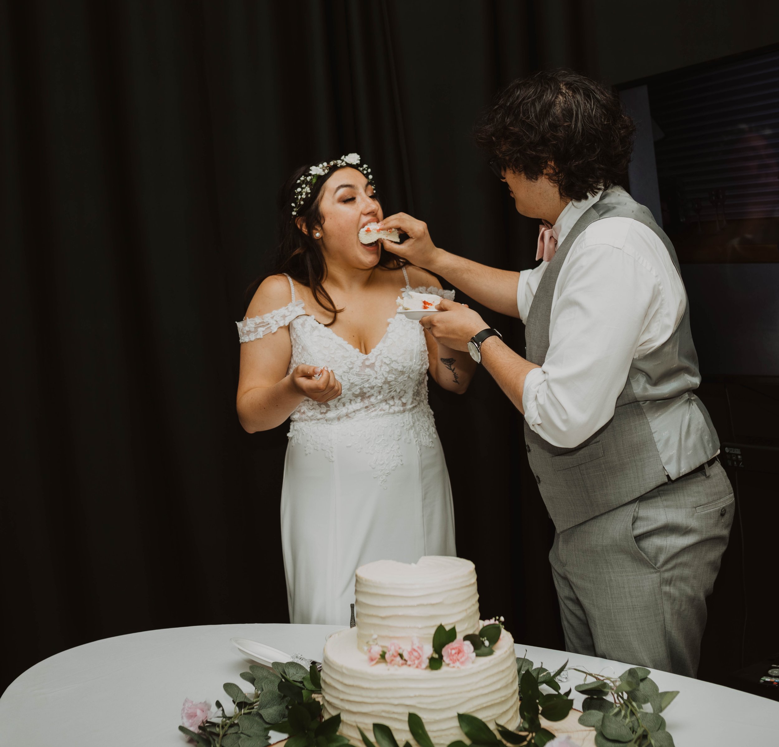 A bride in a white wedding dress receiving cake from a groom in a light gray vest and pants at her wedding celebration. Seattle, WA wedding photography.