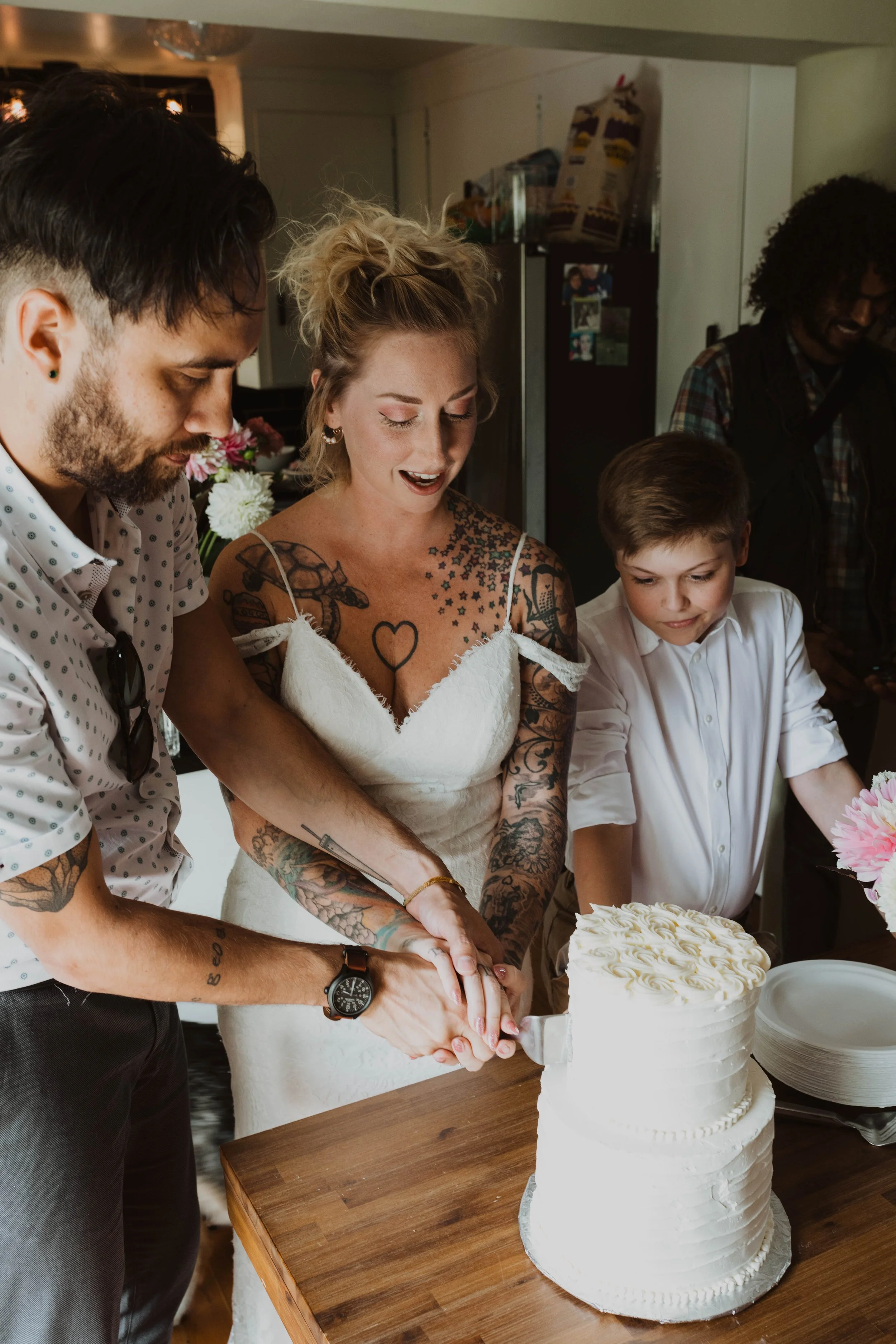 A bride, groom, and young boy cutting a white wedding cake together, with people in the background. Seattle, WA wedding photography.