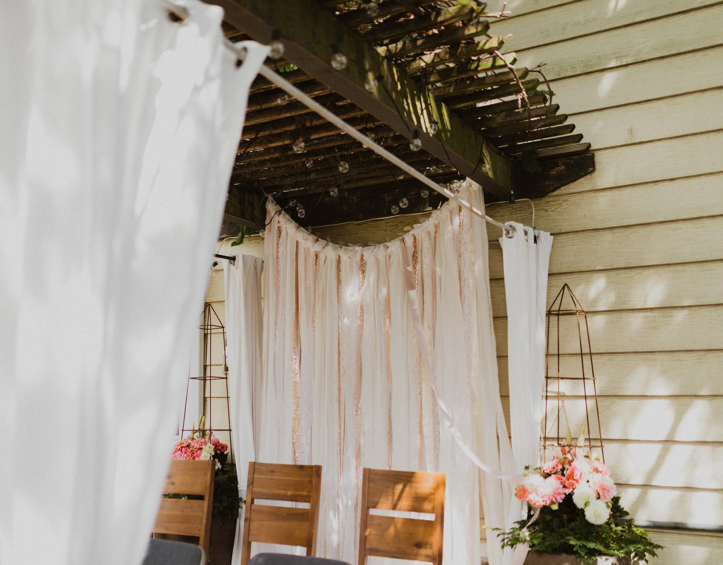Decorative outdoor space with white curtains, wooden chairs, pink and white flowers, and a backdrop of a pink and white fabric curtain, with a wooden wall and a wooden overhang. Seattle, WA wedding photography.