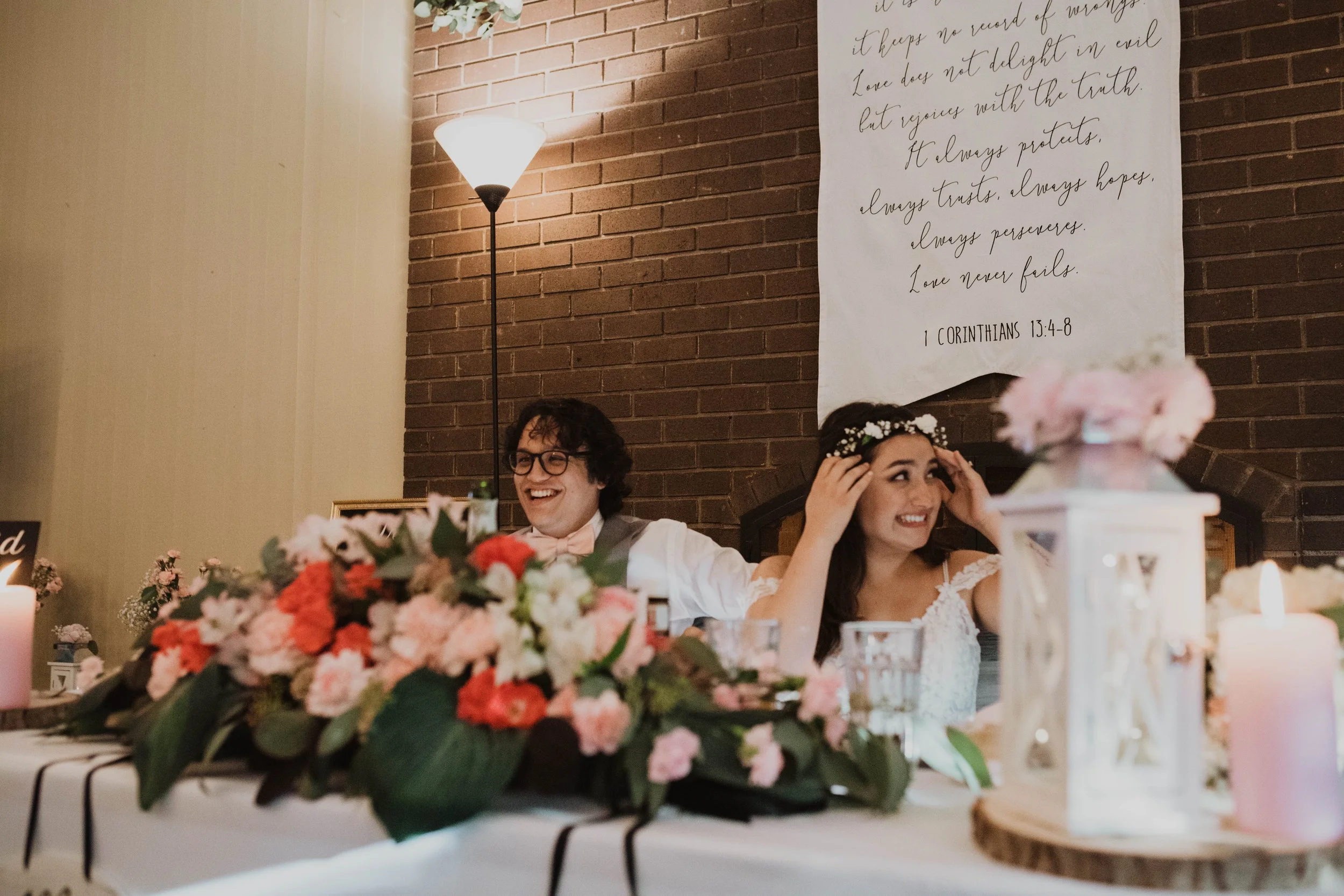 A bride and groom sit at a decorated table during a wedding reception, smiling and laughing. The bride wears a white dress with lace details and a floral headband, while the groom wears glasses, a vest, and a bow tie. The table is adorned with pink, 