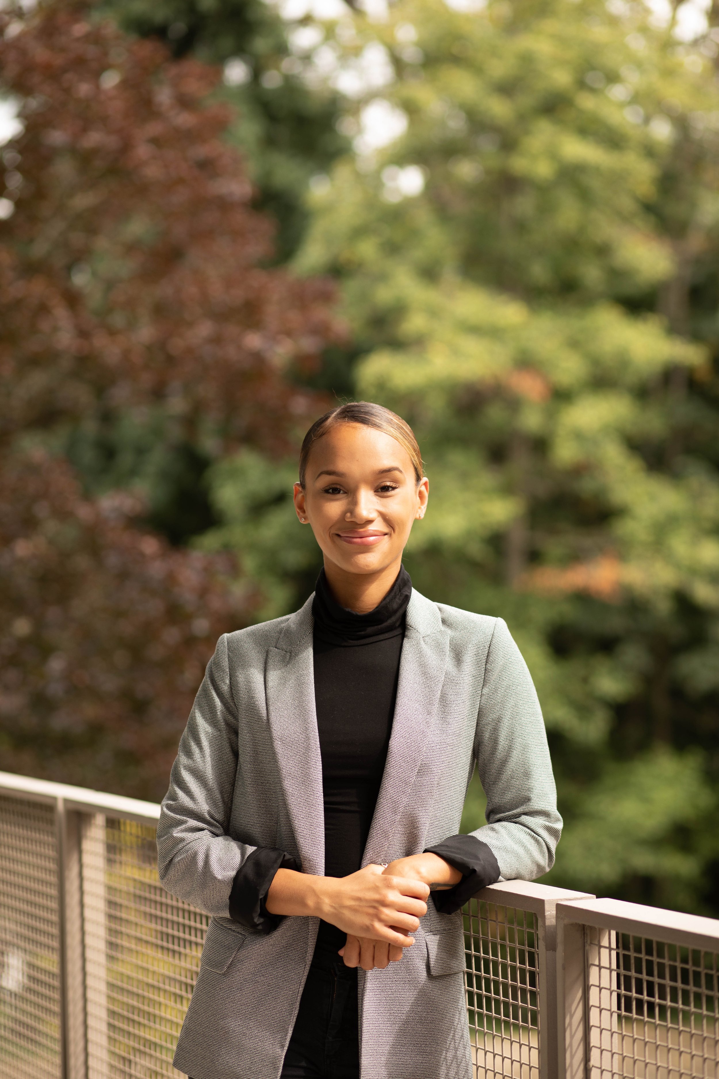 A professional woman in a gray blazer and black turtleneck standing outdoors on a bridge with lush green trees in the background. Seattle professional head shot photography