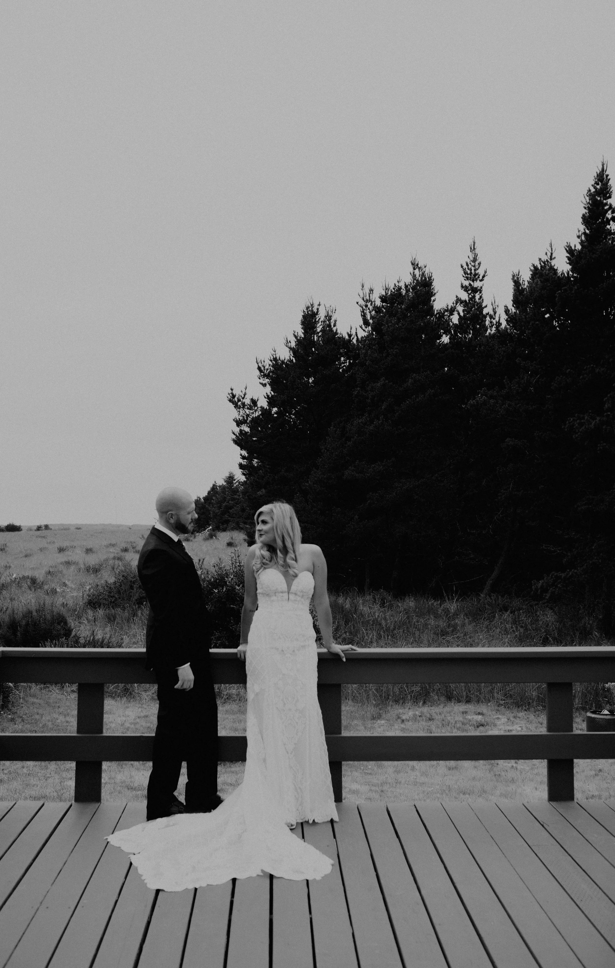 A black and white photograph of a bride and groom standing on a wooden deck, facing each other, with a backdrop of trees and open field. Long Beach, WA wedding photography.