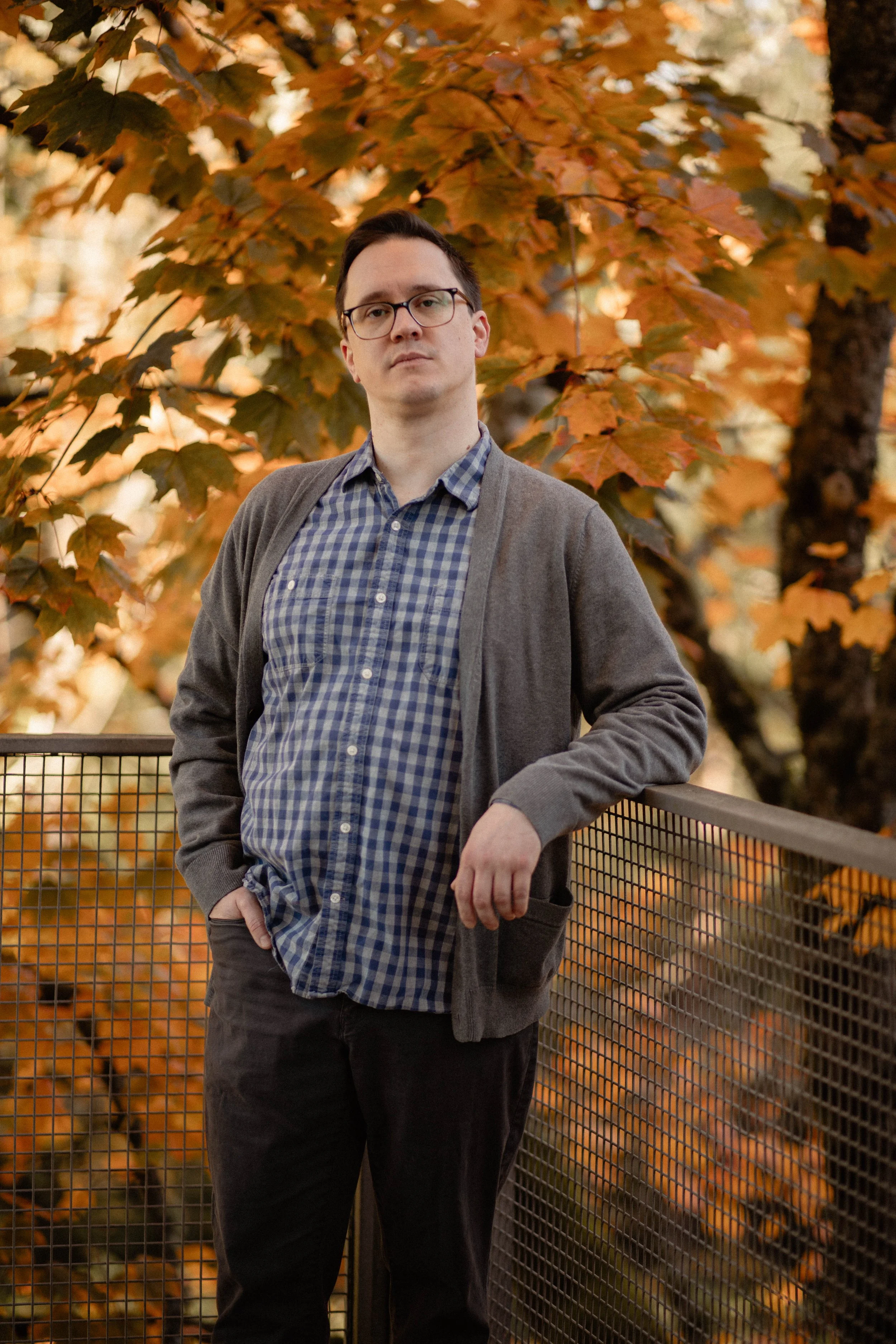 A man in glasses, a checkered shirt, and a gray cardigan leaning on a metal railing with autumn leaves in the background. Seattle professional head shot photography