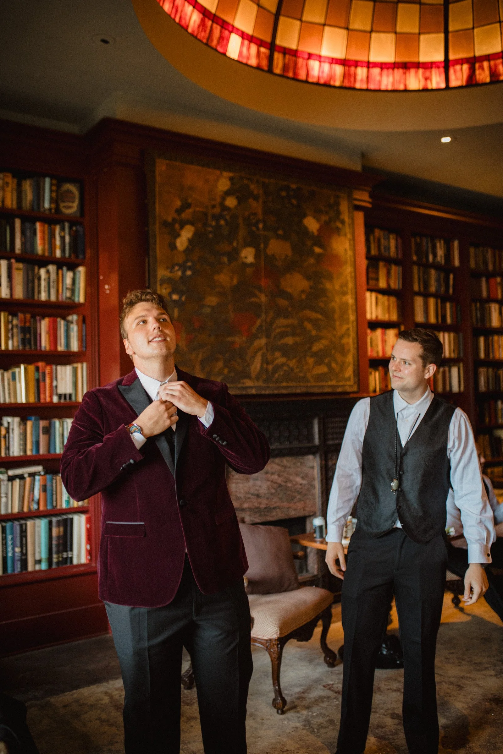 Groom adjusts his tie while his groomsmen stands nearby in support. In the library of The Ruins wedding venue, Queen Anne, Seattle