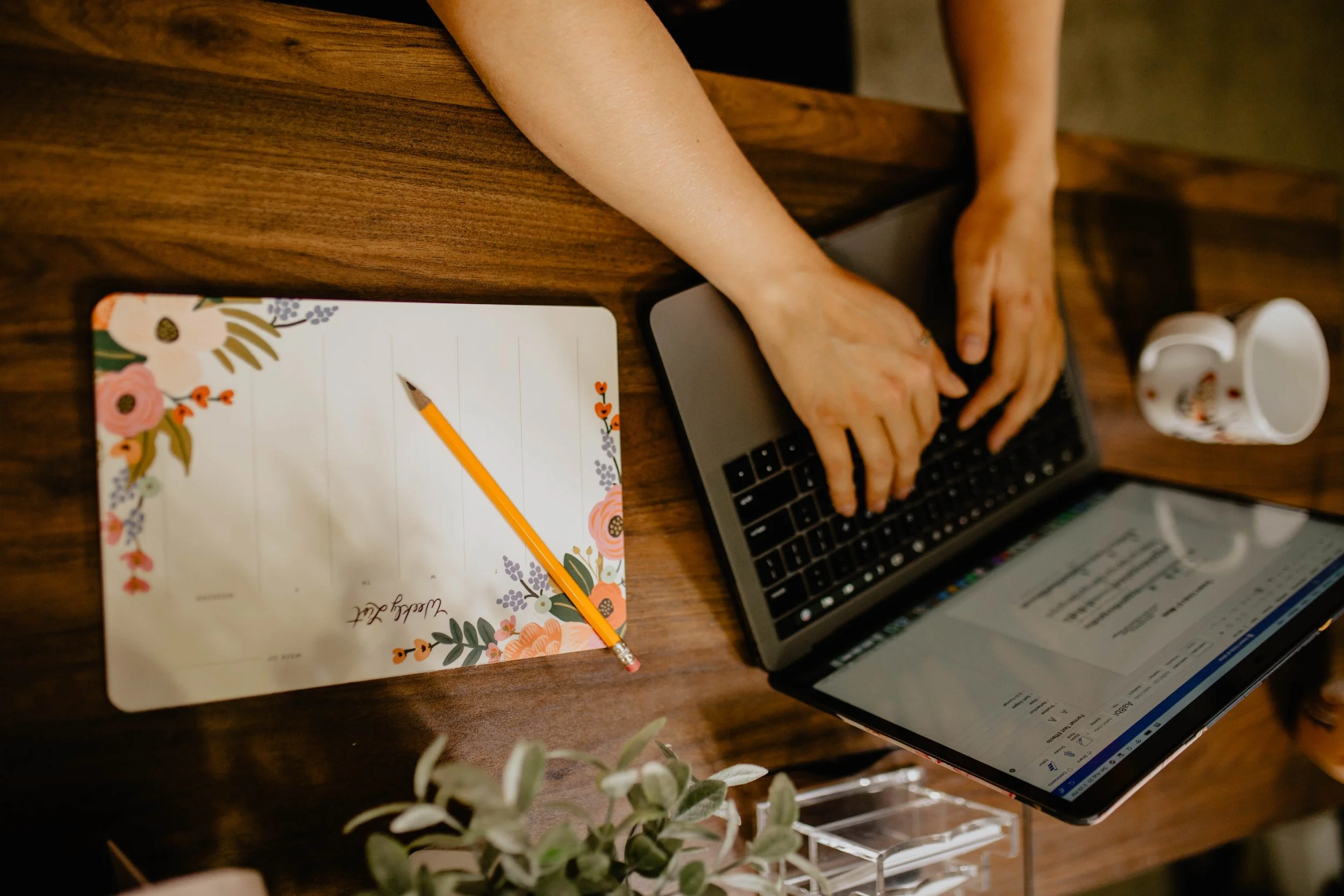 Person typing on a laptop at a wooden desk with a floral-patterned notepad, a pencil, a white mug, and some green leafy plant in the foreground. Seattle professional head shot photography