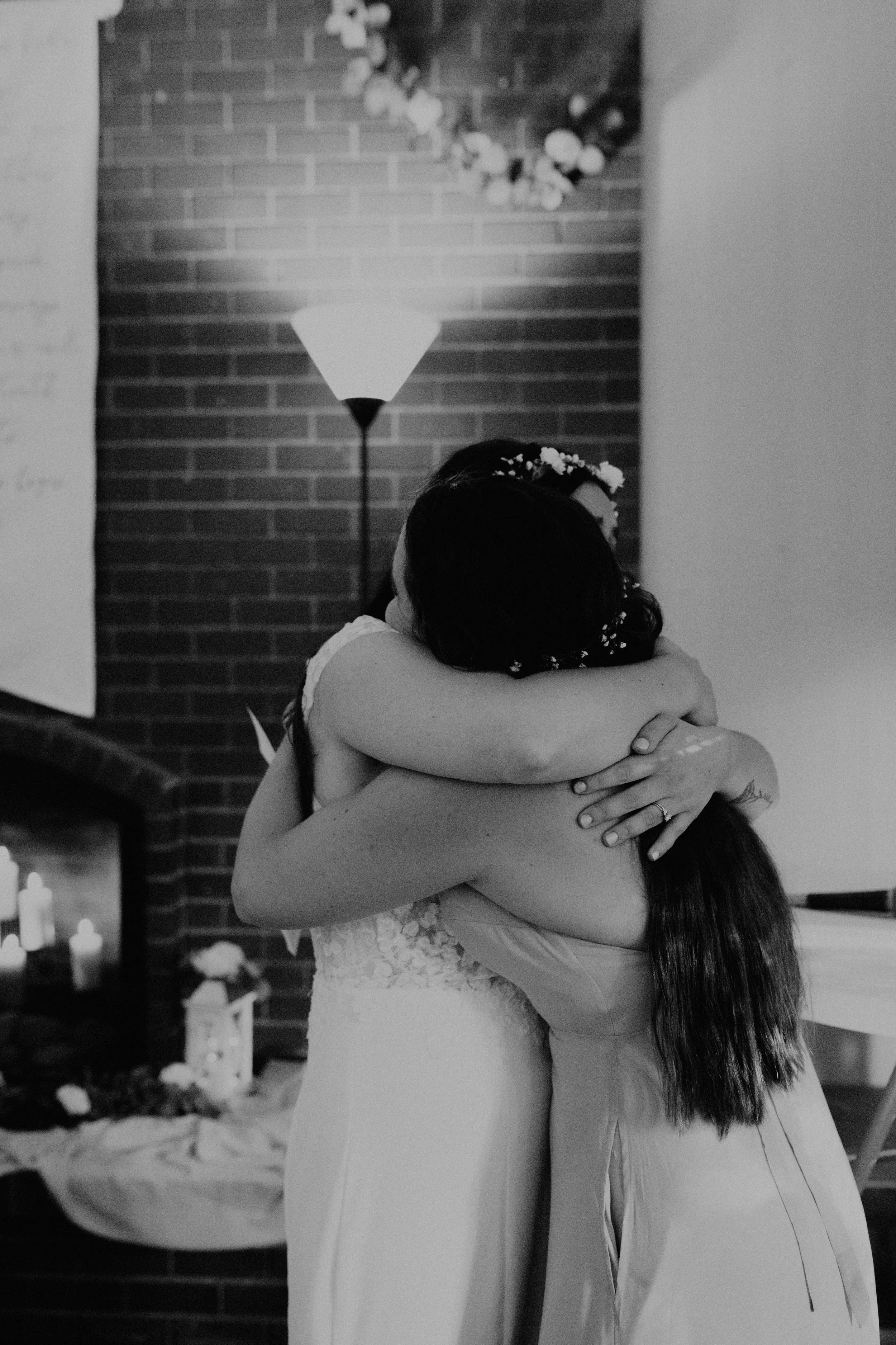 Two women hugging each other at a wedding, with one wearing a wedding dress and the other in a light-colored dress, in front of a brick wall with a lit floor lamp and flowers. Seattle, WA wedding photography.