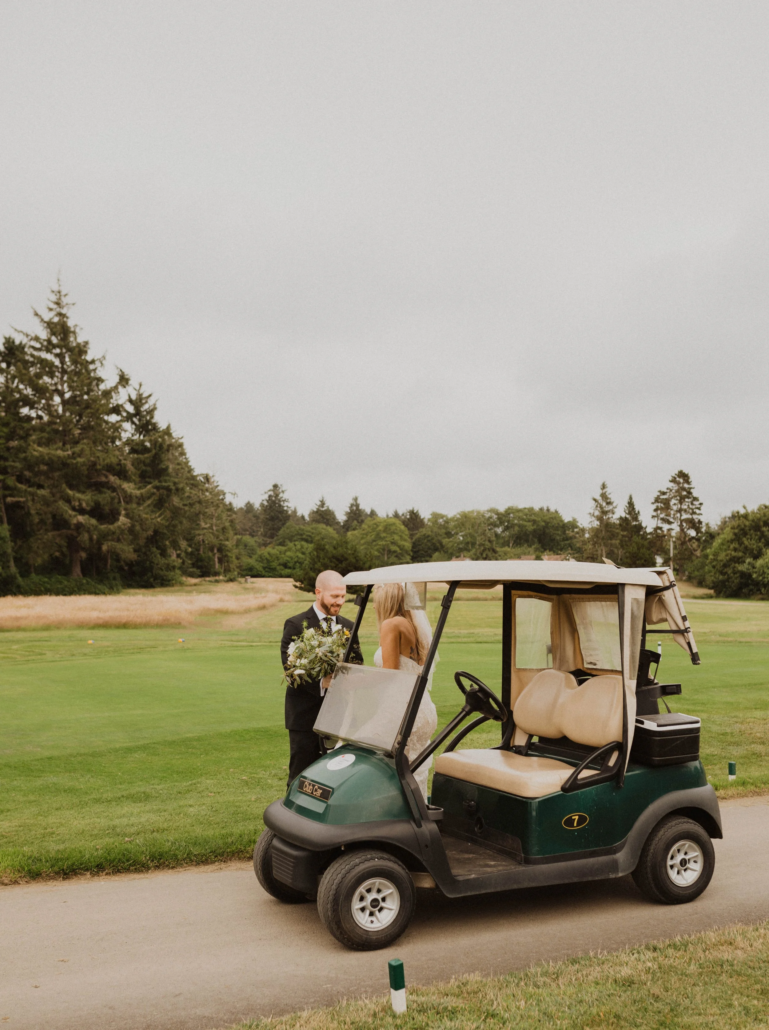 A man in a suit and a woman in a wedding dress standing by a golf cart on a golf course with greenery and trees in the background. Long Beach, WA wedding photography.