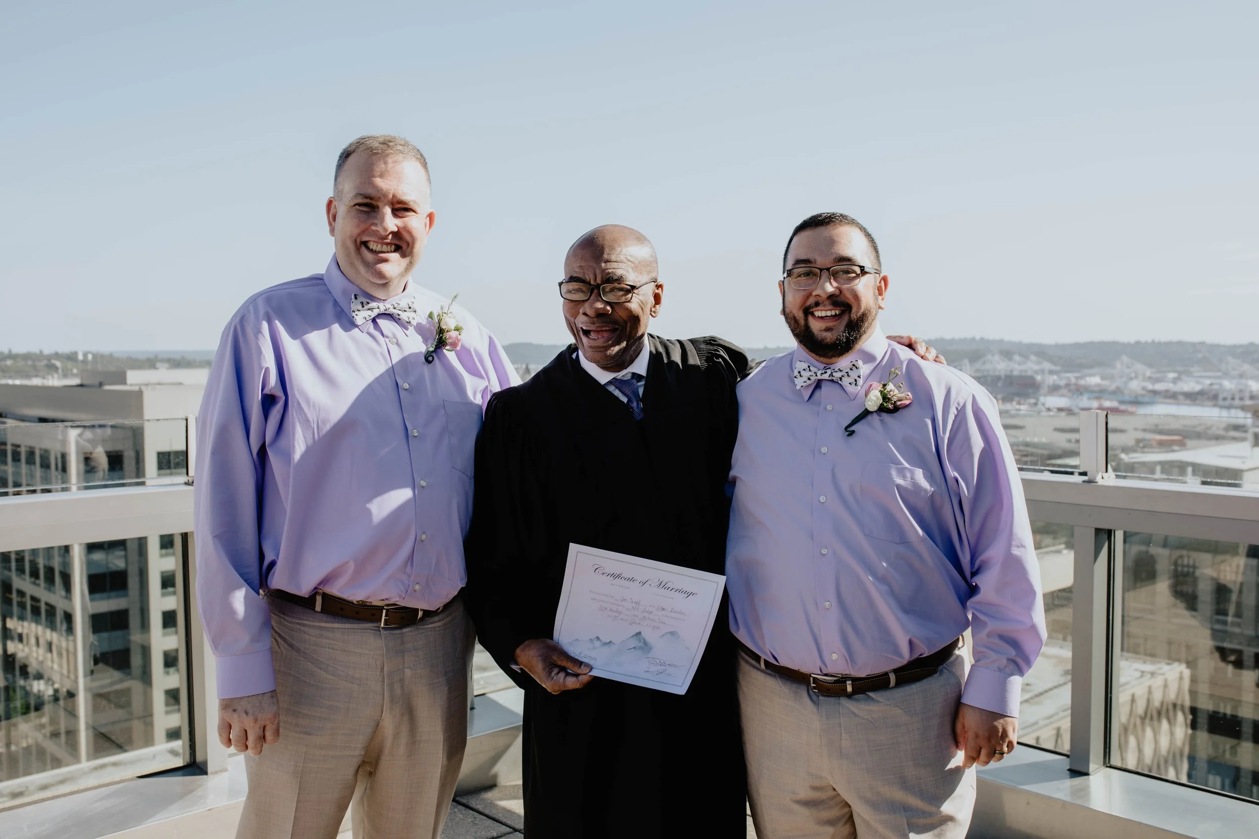 Three men at a wedding celebration on a rooftop terrace, with city buildings and water in the background. The man in the middle wears a black robe and holds a marriage certificate. The two men on either side wear light purple shirts, matching bow tie