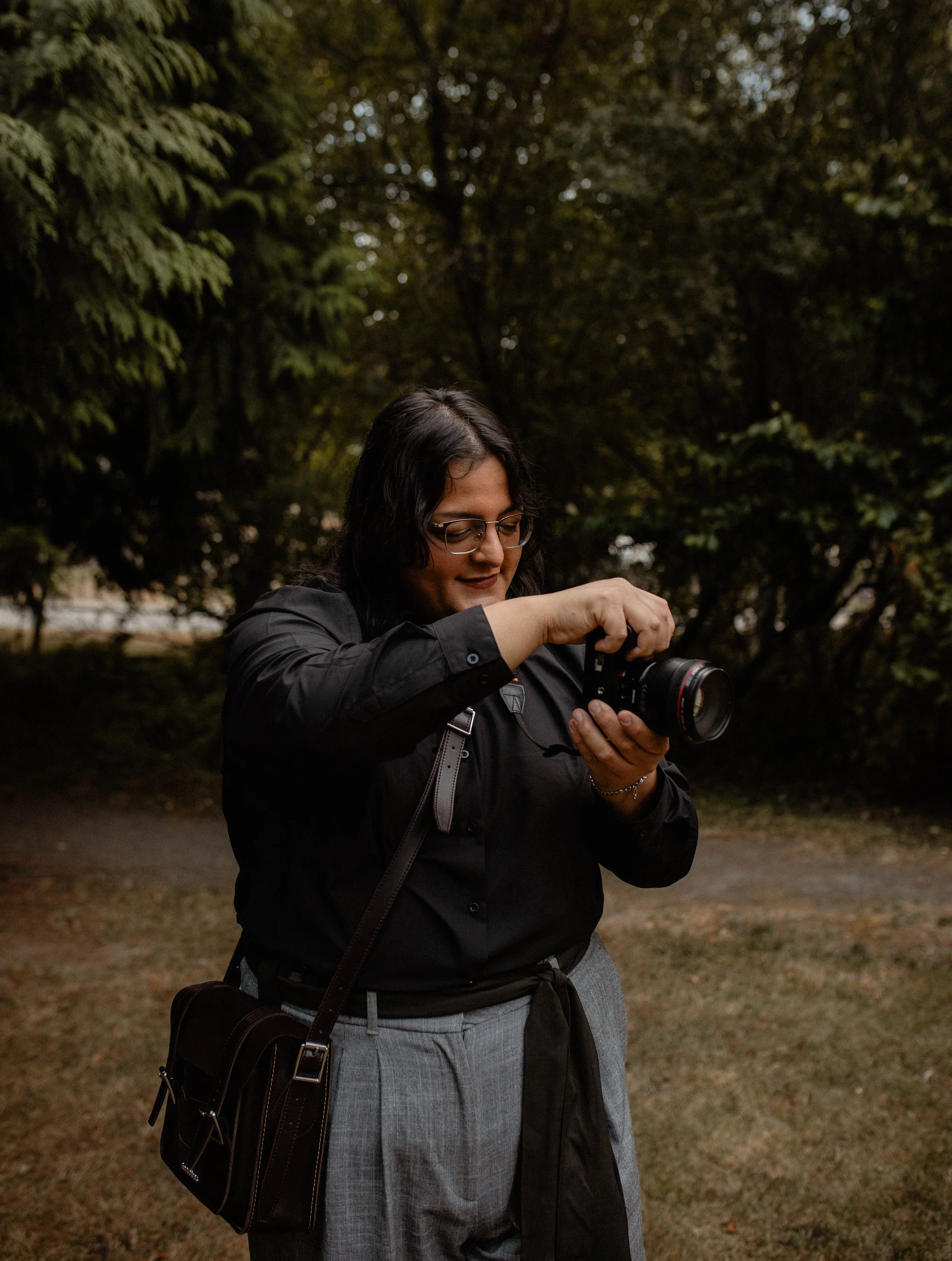 A woman adjusting a camera outdoors in a park or wooded area, wearing glasses and a dark shirt, with trees in the background. Seattle professional head shot photography