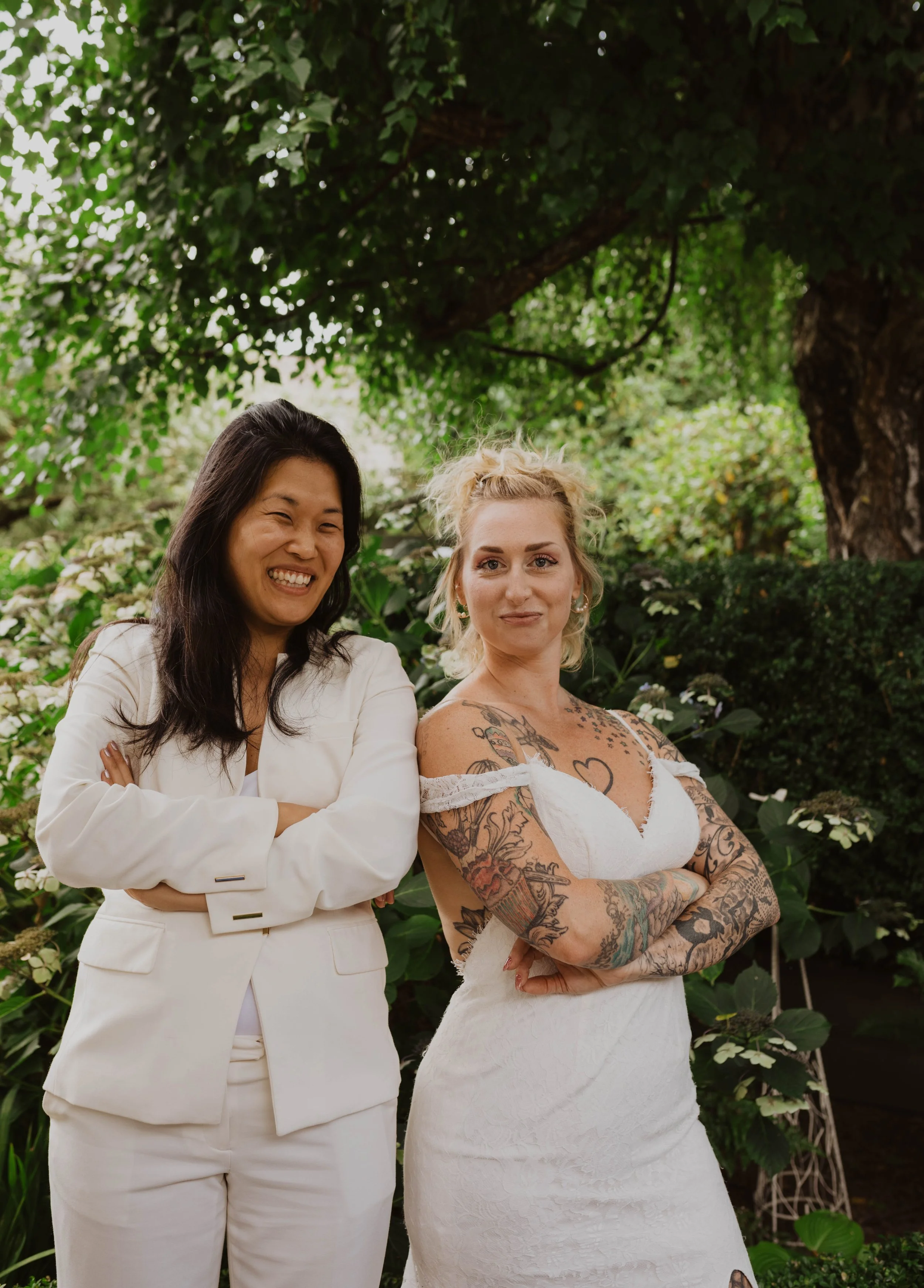 Two women standing outdoors with green foliage and a tree in the background. One woman has long black hair and is dressed in a white suit, smiling with her arms crossed. The other has blonde hair styled up, tattoos on her arms and chest, and is weari