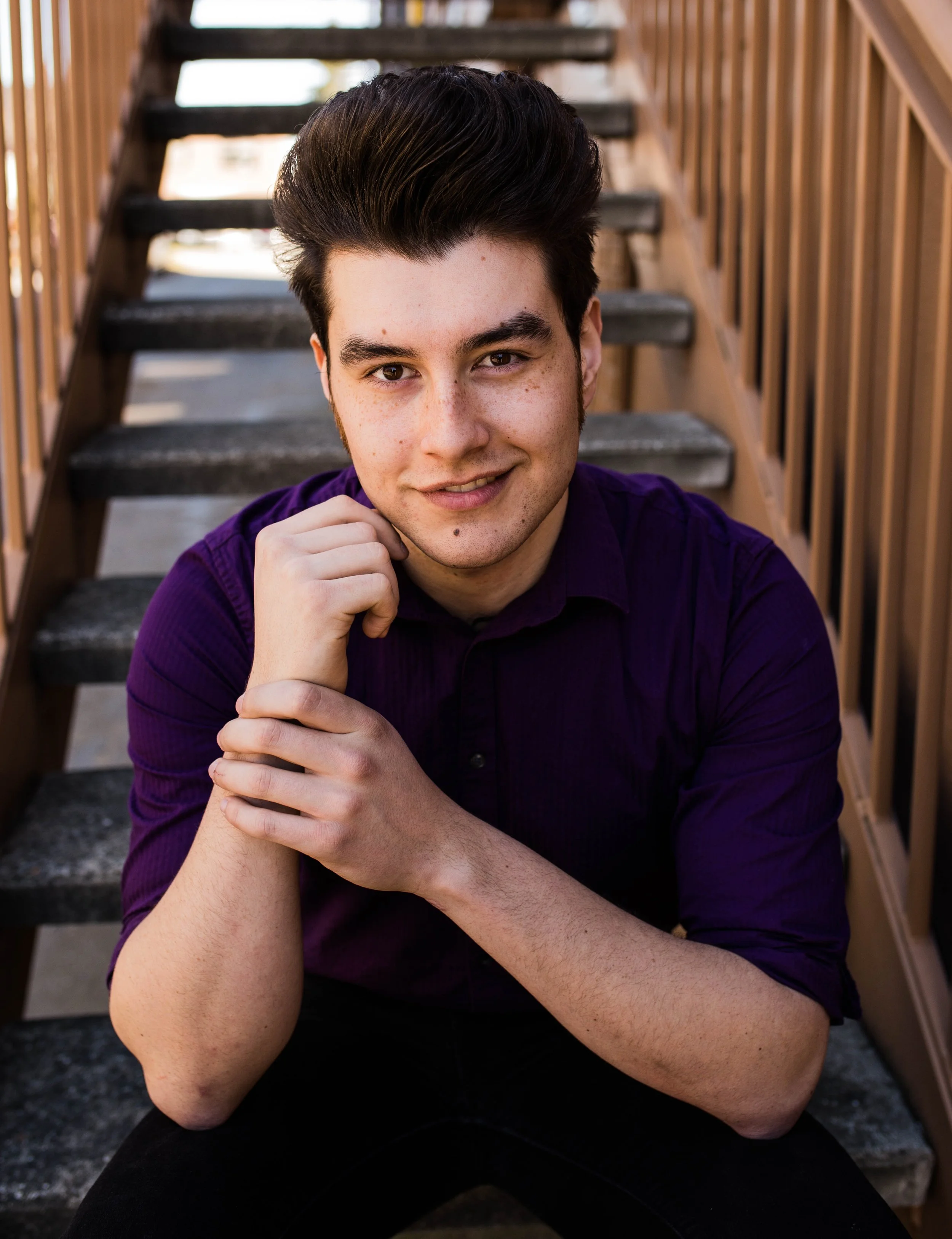 Young man with dark hair and light skin sitting on outdoor stairs, wearing a purple shirt, smiling at the camera. Seattle professional head shot photography