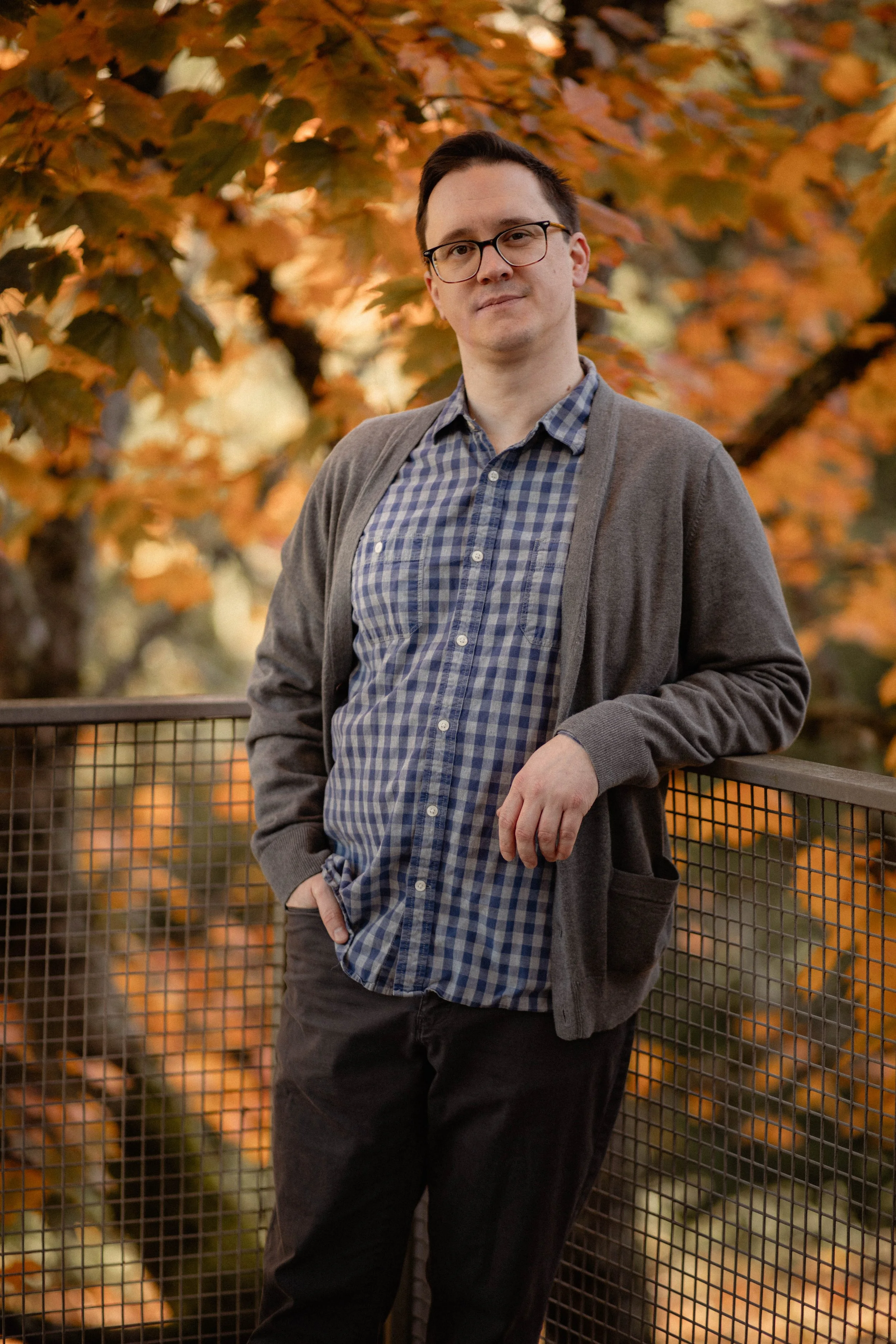 A man wearing glasses, a checkered shirt, and a gray jacket standing outdoors on a railing with autumn leaves in the background. Seattle professional head shot photography