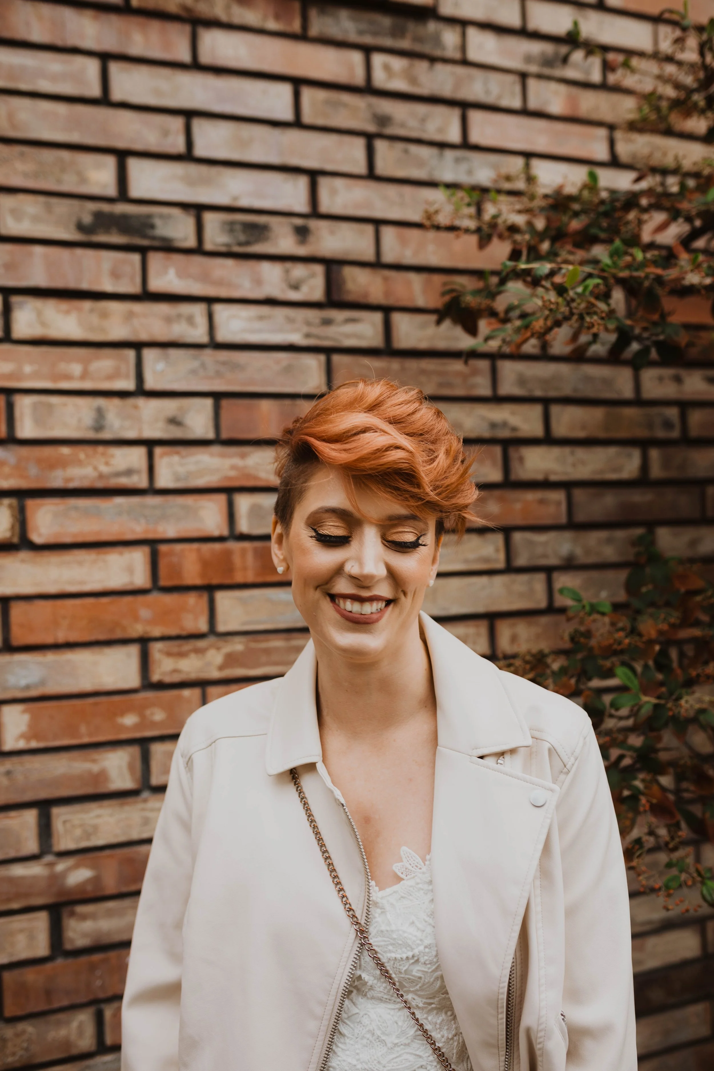 Smiling woman with short, reddish hair standing in front of a brick wall with some greenery. Pioneer Square, Seattle, WA wedding photography.