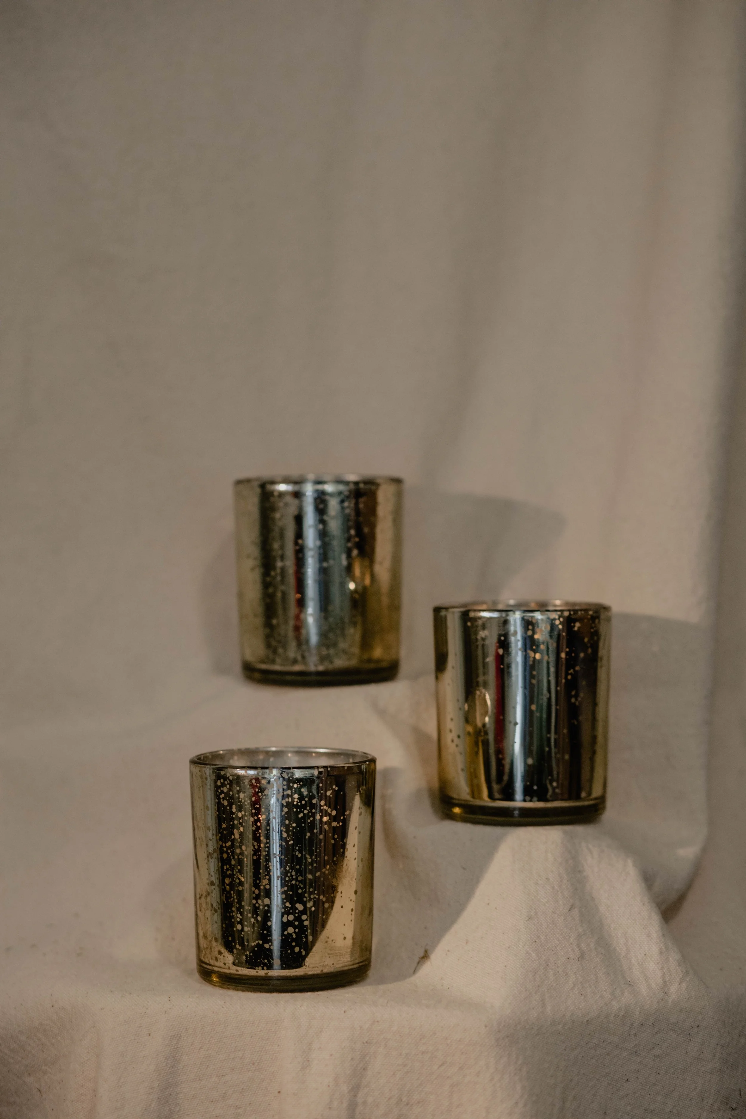 Three silver metallic candles with a speckled pattern on a beige cloth backdrop, arranged with two on the lower level and one on the upper level.  Seattle product photography