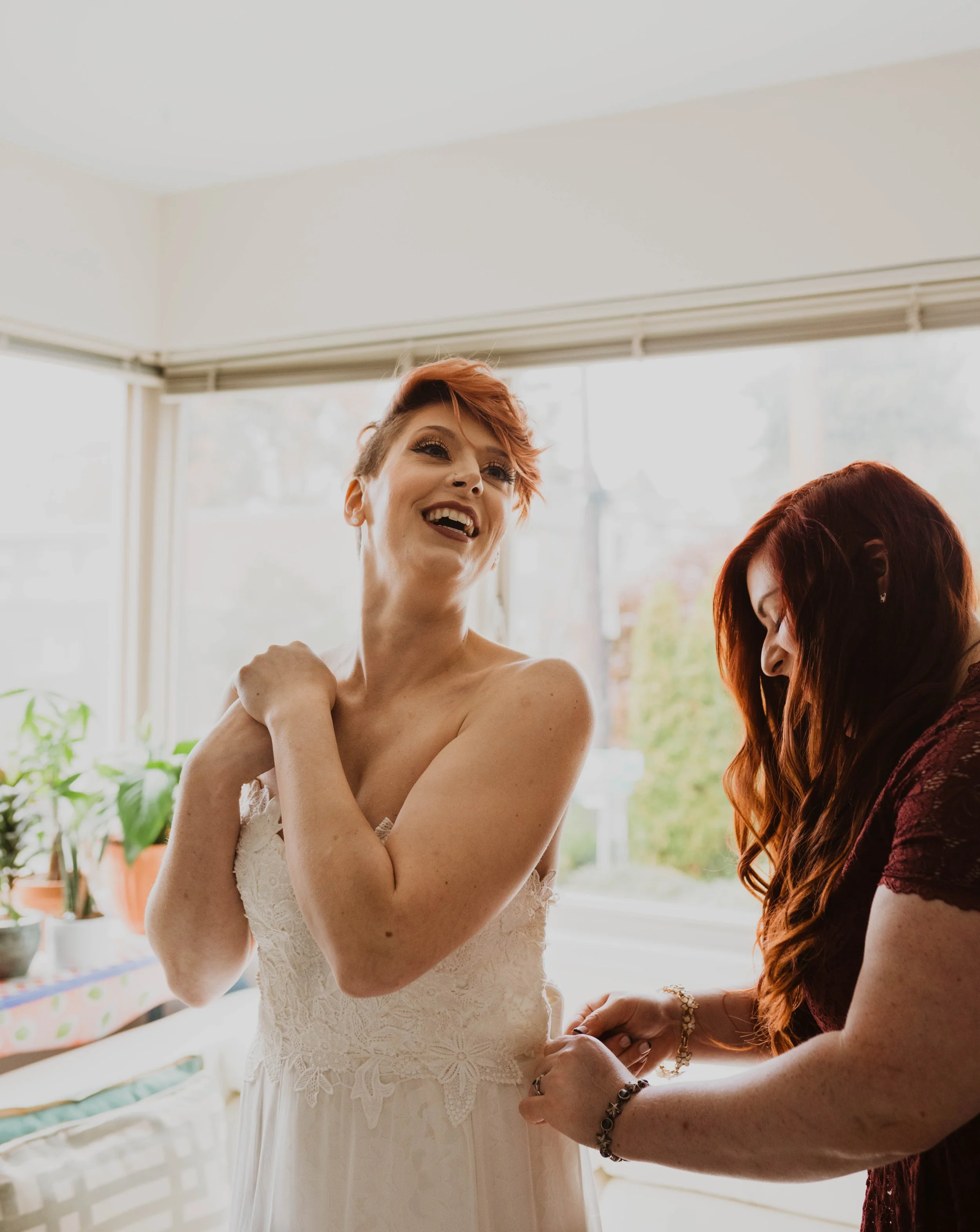 A woman in a white lace dress is smiling and looking up, with another woman in a burgundy dress helping her with her dress near a window. Pioneer Square, Seattle, WA wedding photography.