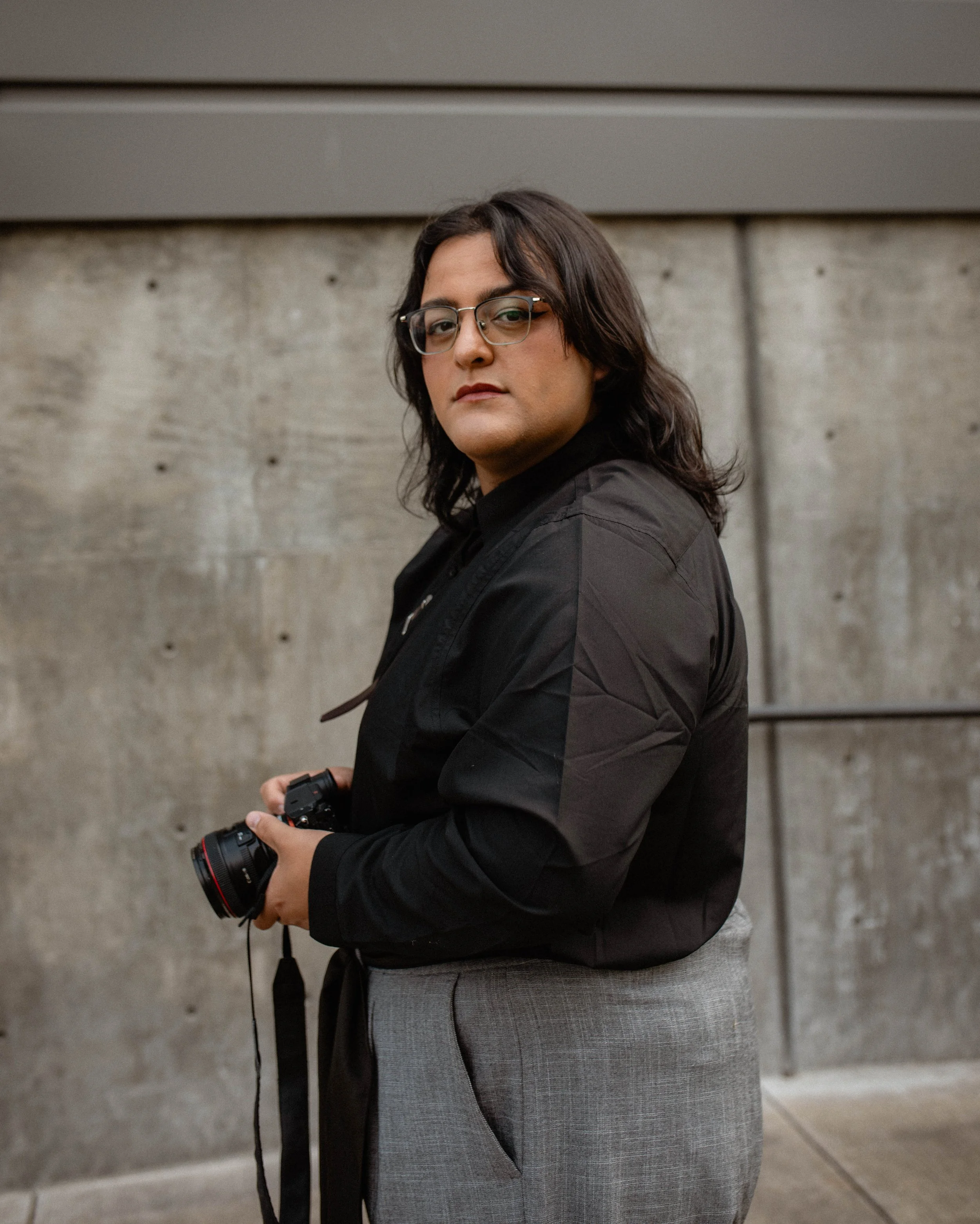 A person with shoulder-length dark hair, glasses, and a serious expression, holding a camera in their hand, standing against a concrete wall. Seattle professional head shot photography