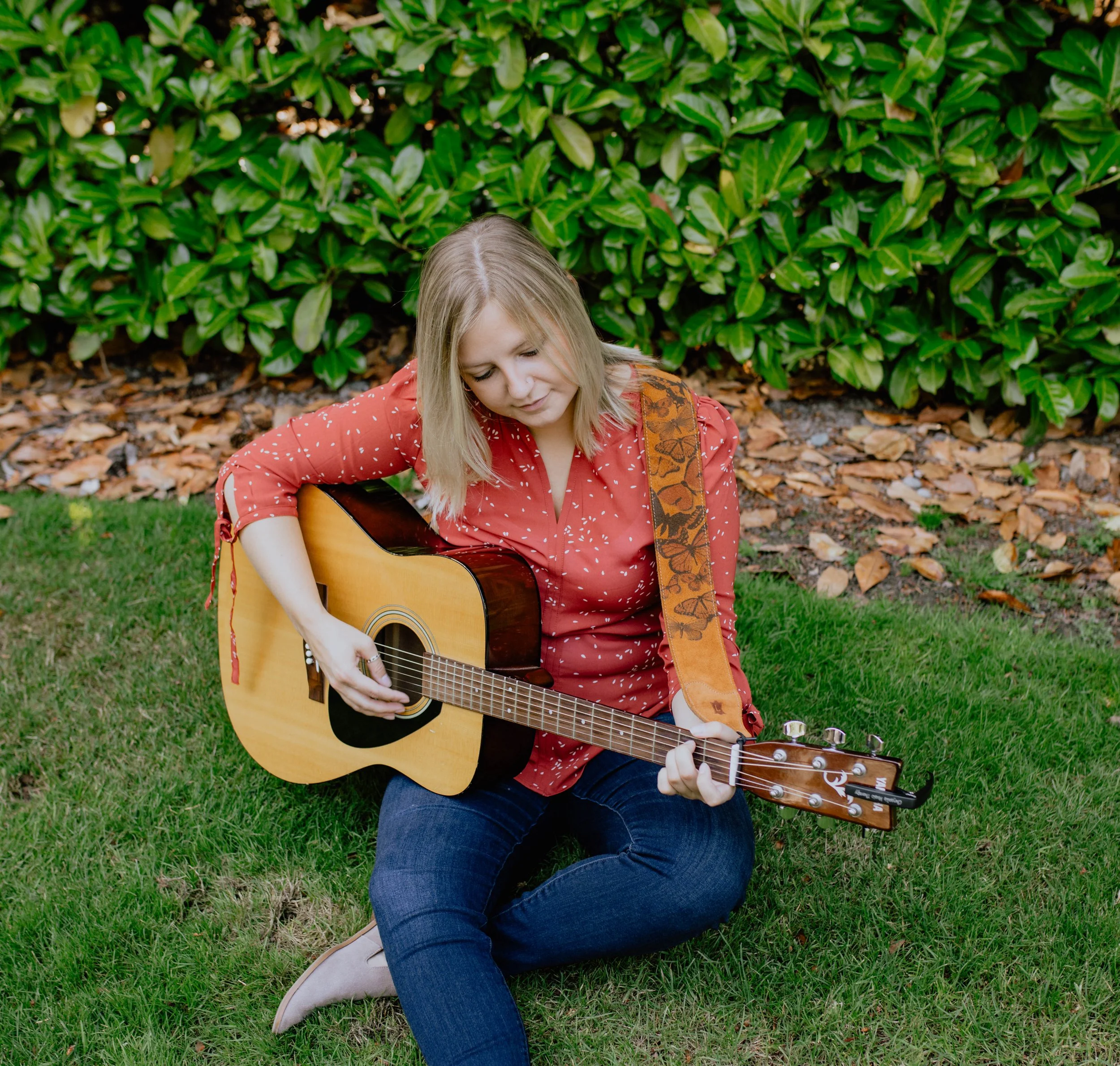 A woman sitting on grass playing an acoustic guitar, with a bush and fallen leaves in the background. Seattle professional head shot photography