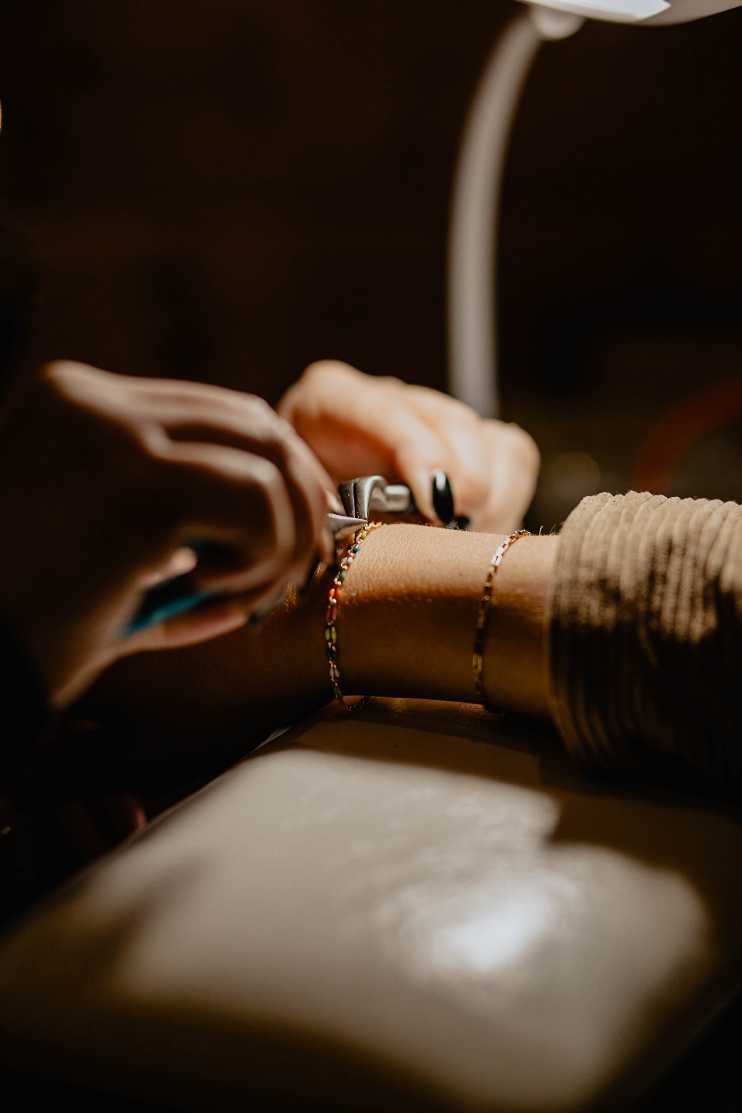 A person is using pliers to attach jewelry, possibly a bracelet or necklace, on their wrist. The person has dark nail polish and is wearing a beige or brown long-sleeved top. The focus is on their hands and the jewelry work, with a blurred dark backg