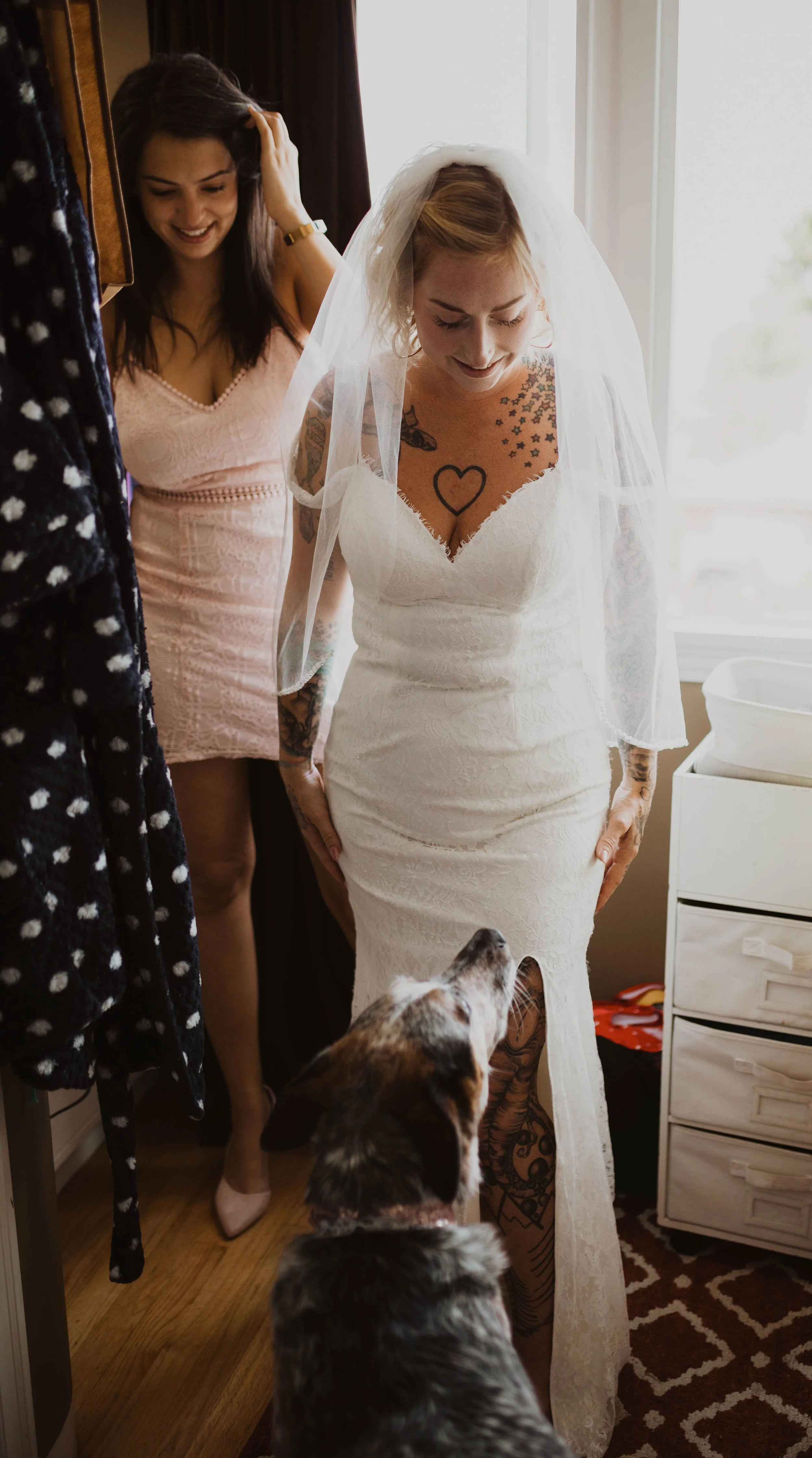 Bride in a white wedding dress and veil smiling at a dog, with a woman in a pink dress behind her, in a room with natural light. Seattle, WA wedding photography.