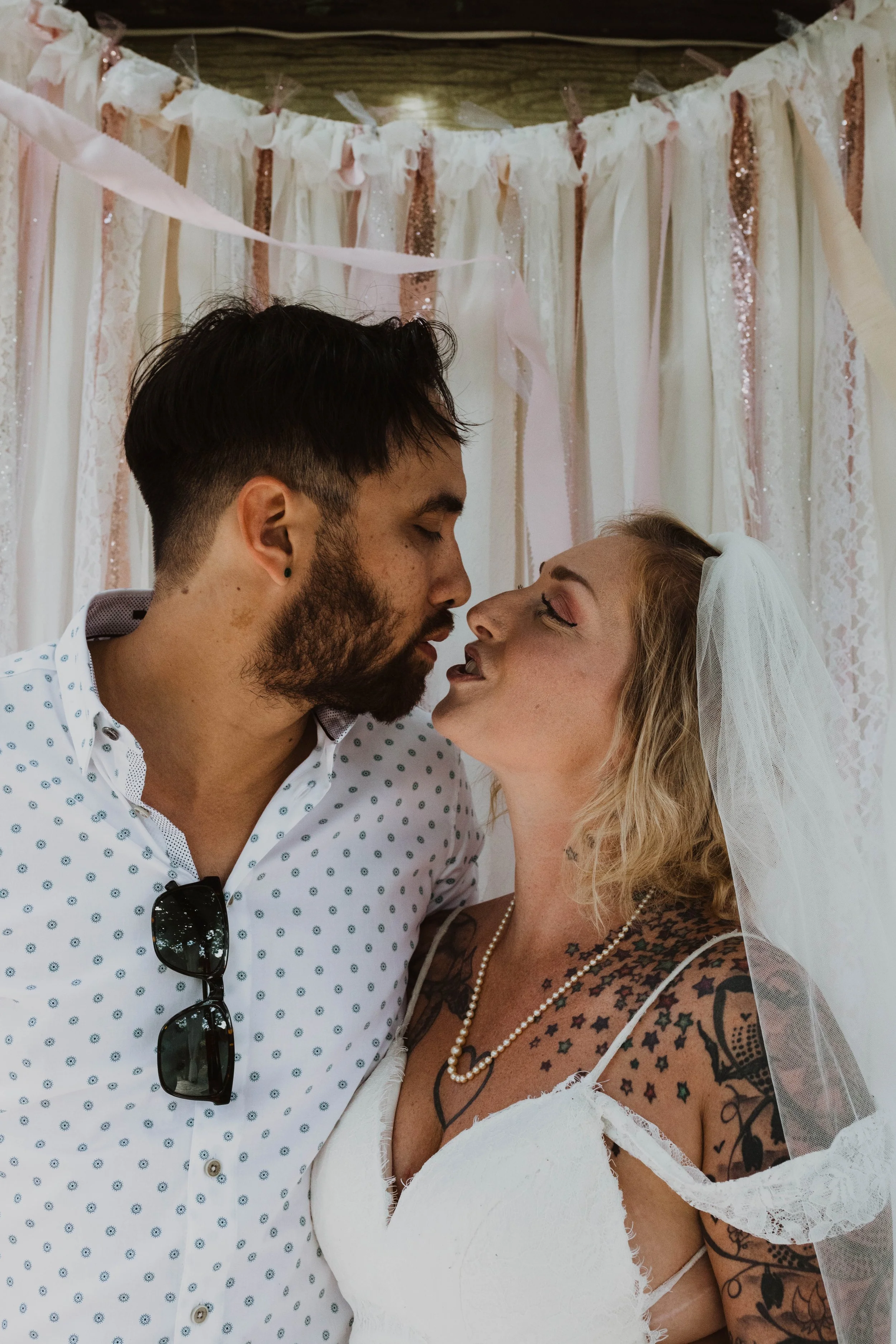 A couple with closed eyes and slightly parted lips, about to kiss, standing in front of a pink and white decorative backdrop, with the woman wearing a veil and the man wearing sunglasses hanging from his shirt. Seattle, WA wedding photography.