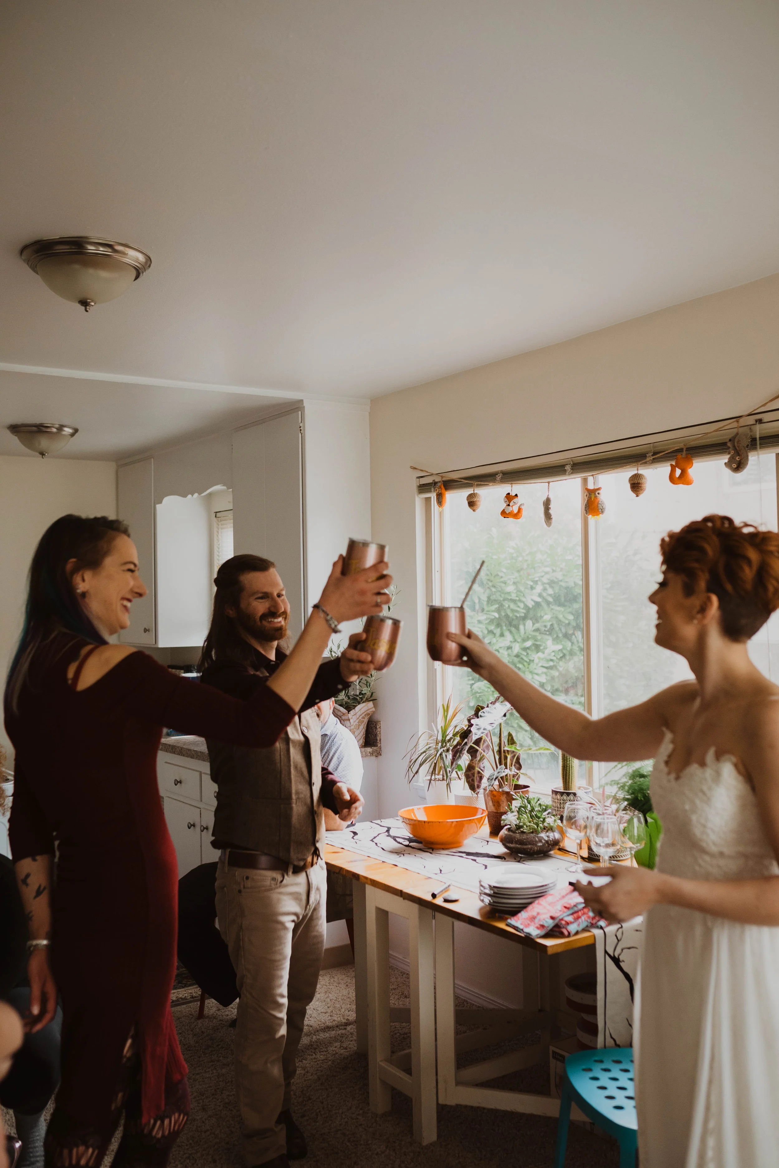 People laughing and clinking cans with straws in a cozy kitchen during a celebration, with plants and decorations in the background. Pioneer Square, Seattle, WA wedding photography.