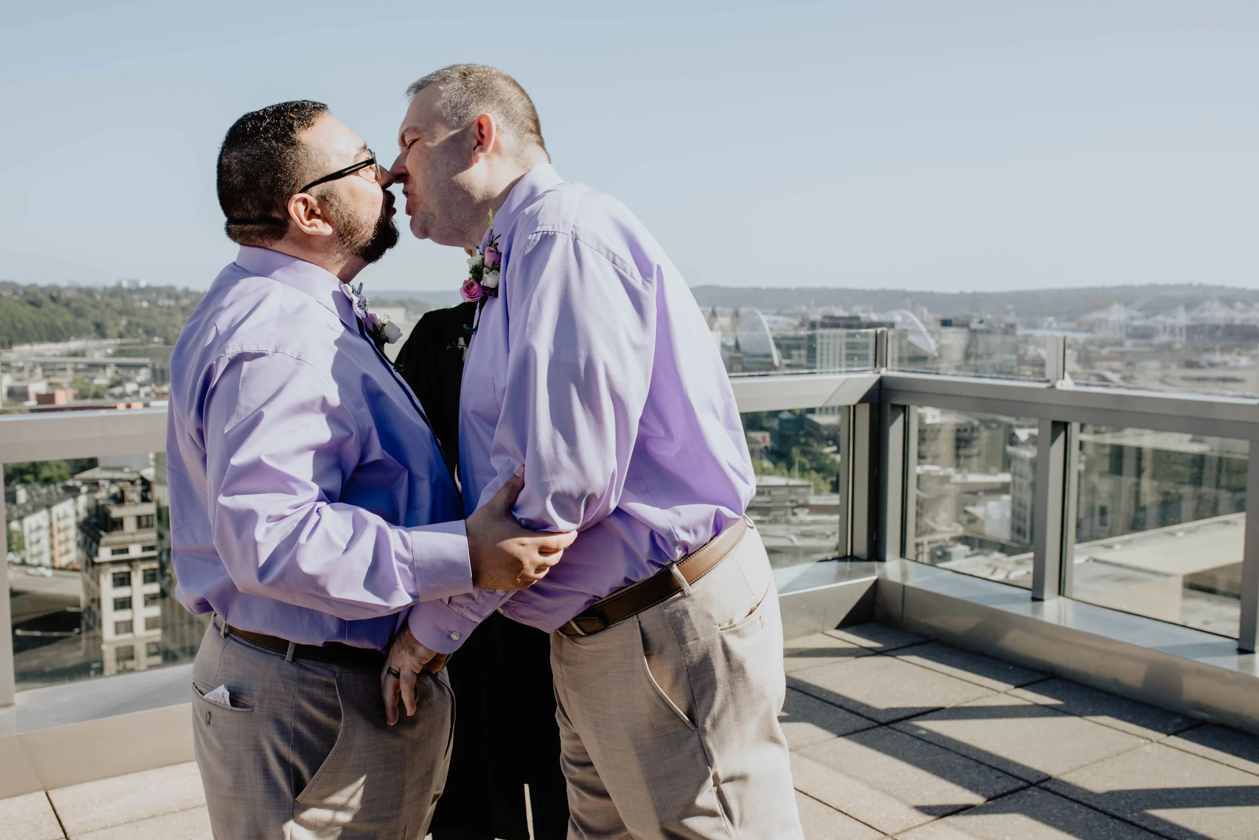 Two men in light purple shirts and beige pants sharing a kiss on a rooftop with cityscape in the background. Seattle Municipal Courthouse wedding photography.