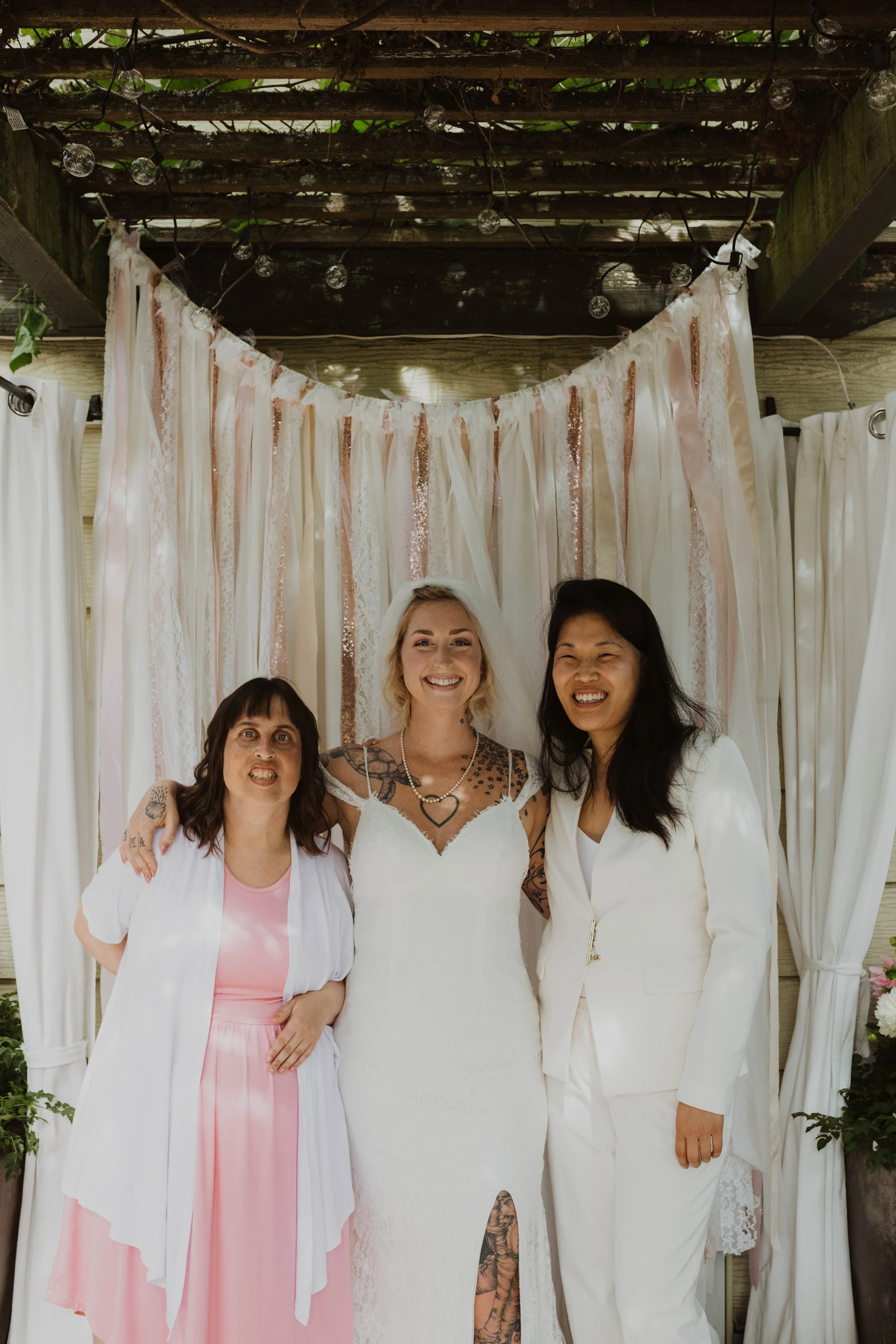 Three women smiling and posing together at a wedding venue, with a backdrop of tissue paper and fabric decorations, and string lights overhead. Seattle, WA wedding photography.