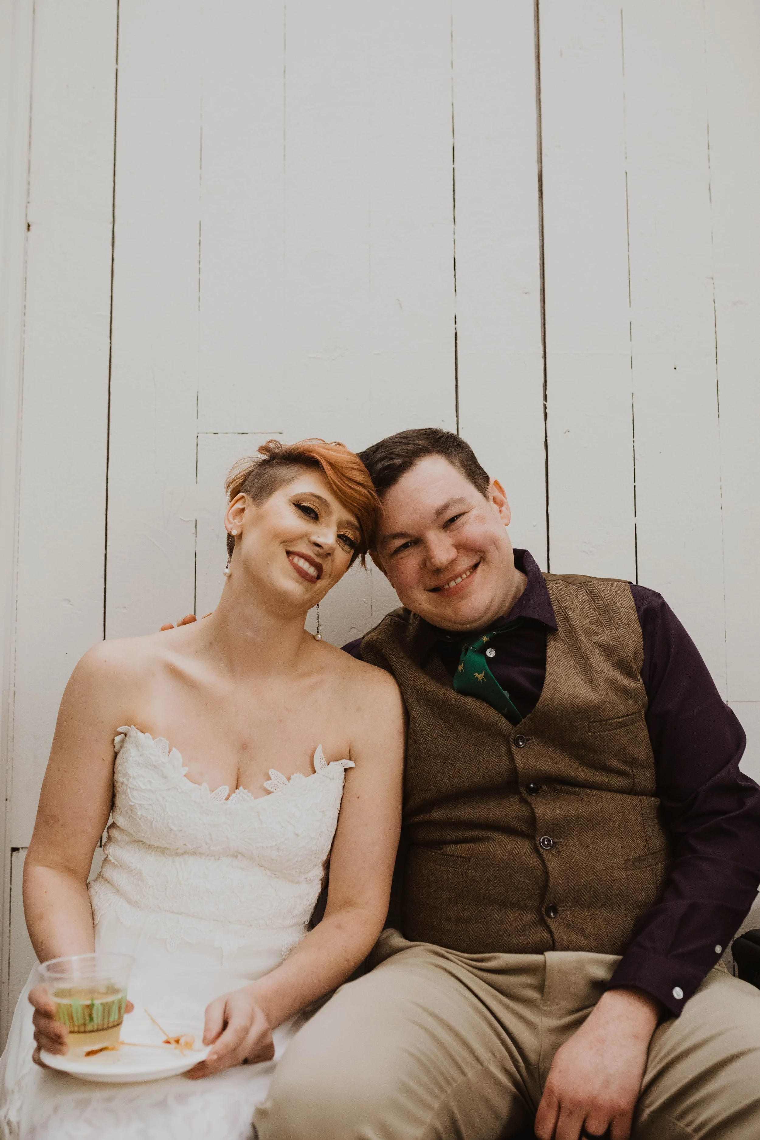 A smiling woman in a white wedding dress sitting next to a smiling man in a brown vest and dark shirt, with a white wooden wall in the background. Pioneer Square, Seattle, WA wedding photography.
