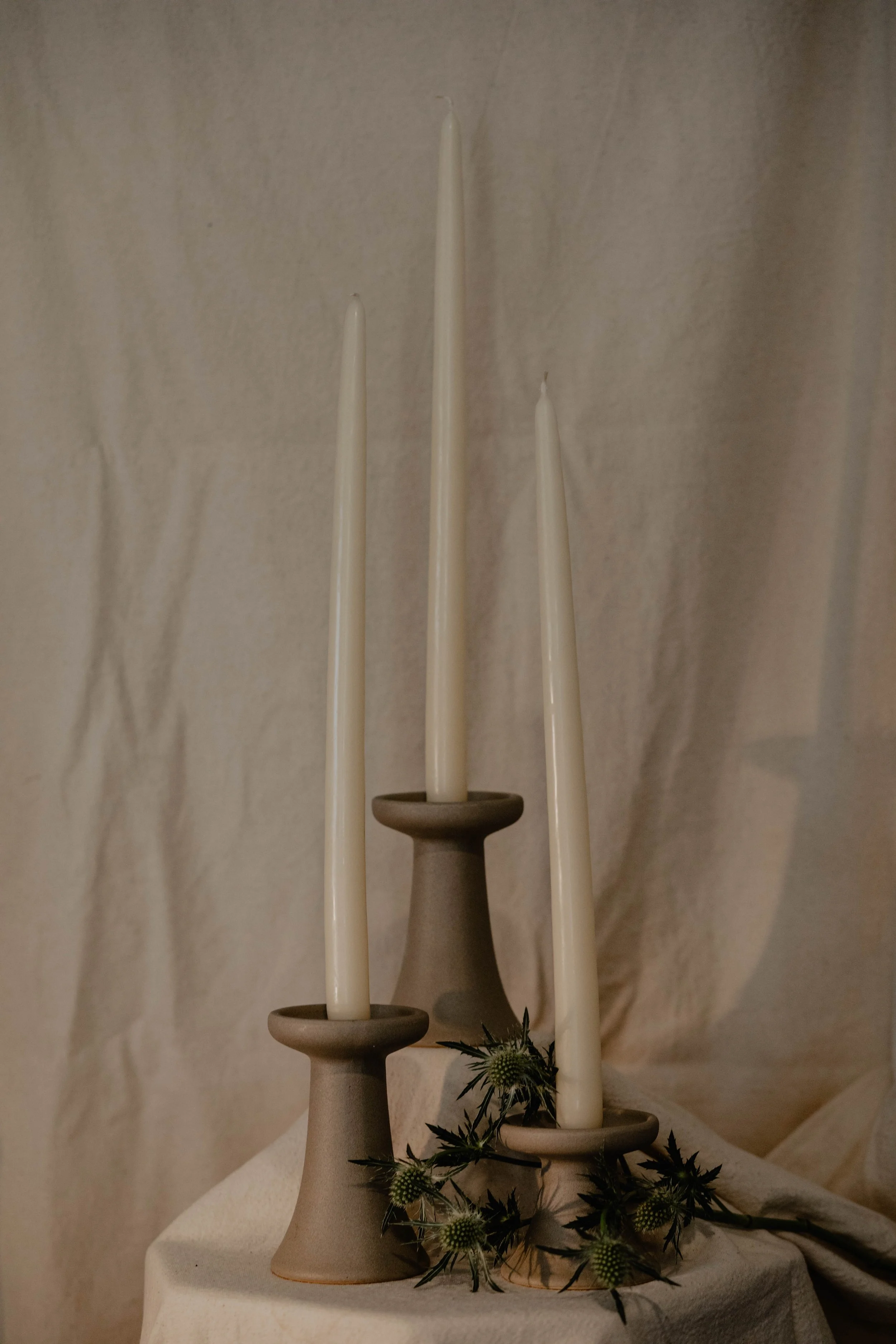 Three beige candles on ceramic holders of varying heights placed on a table with decorative greenery in front of a textured off-white wall.  Seattle product photography