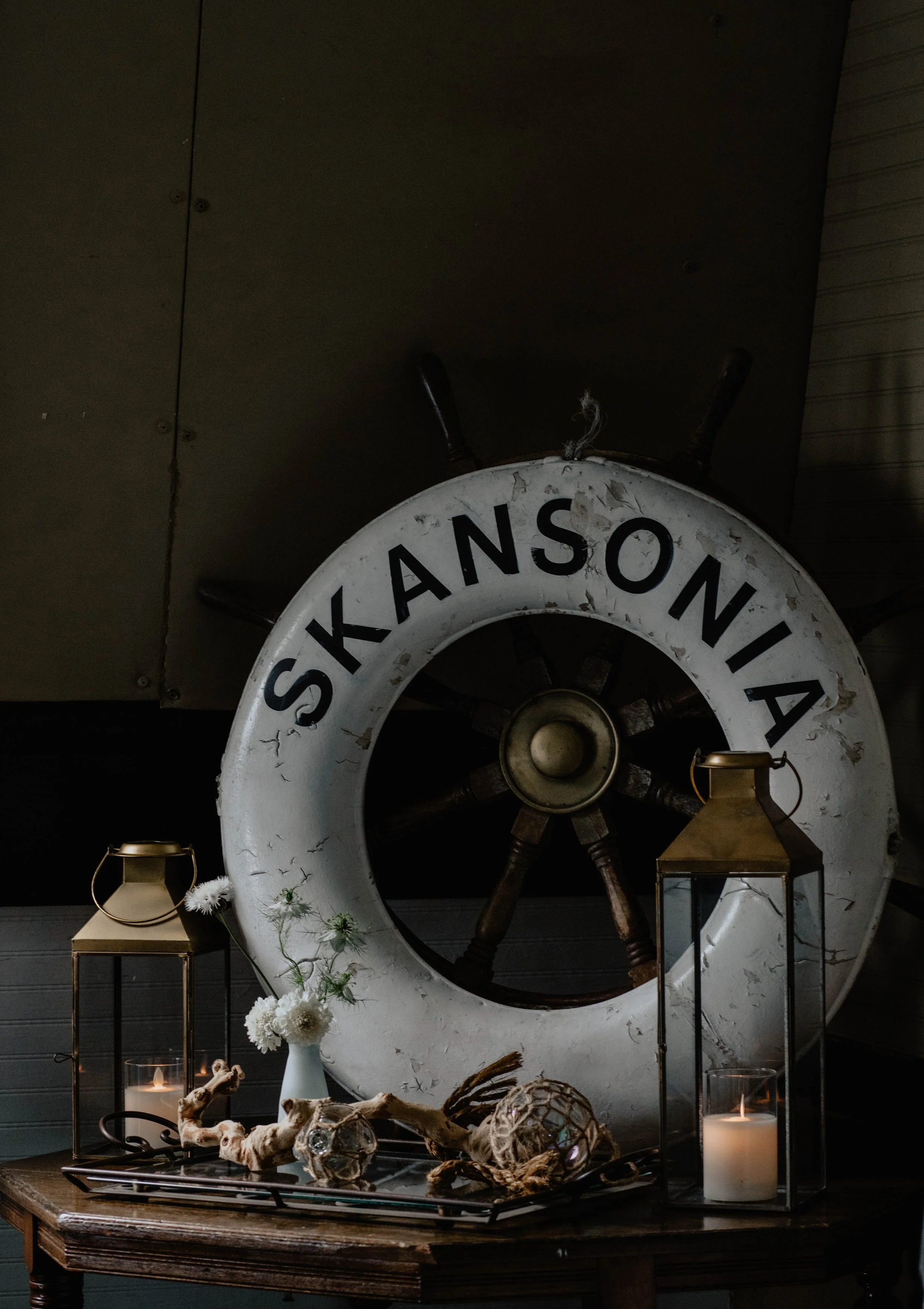 Decorative nautical display with a large ship wheel and a life preserver that has the word 'SKANSONIA' written on it, surrounded by candles, flowers, and driftwood on a wooden table. Seattle event photography