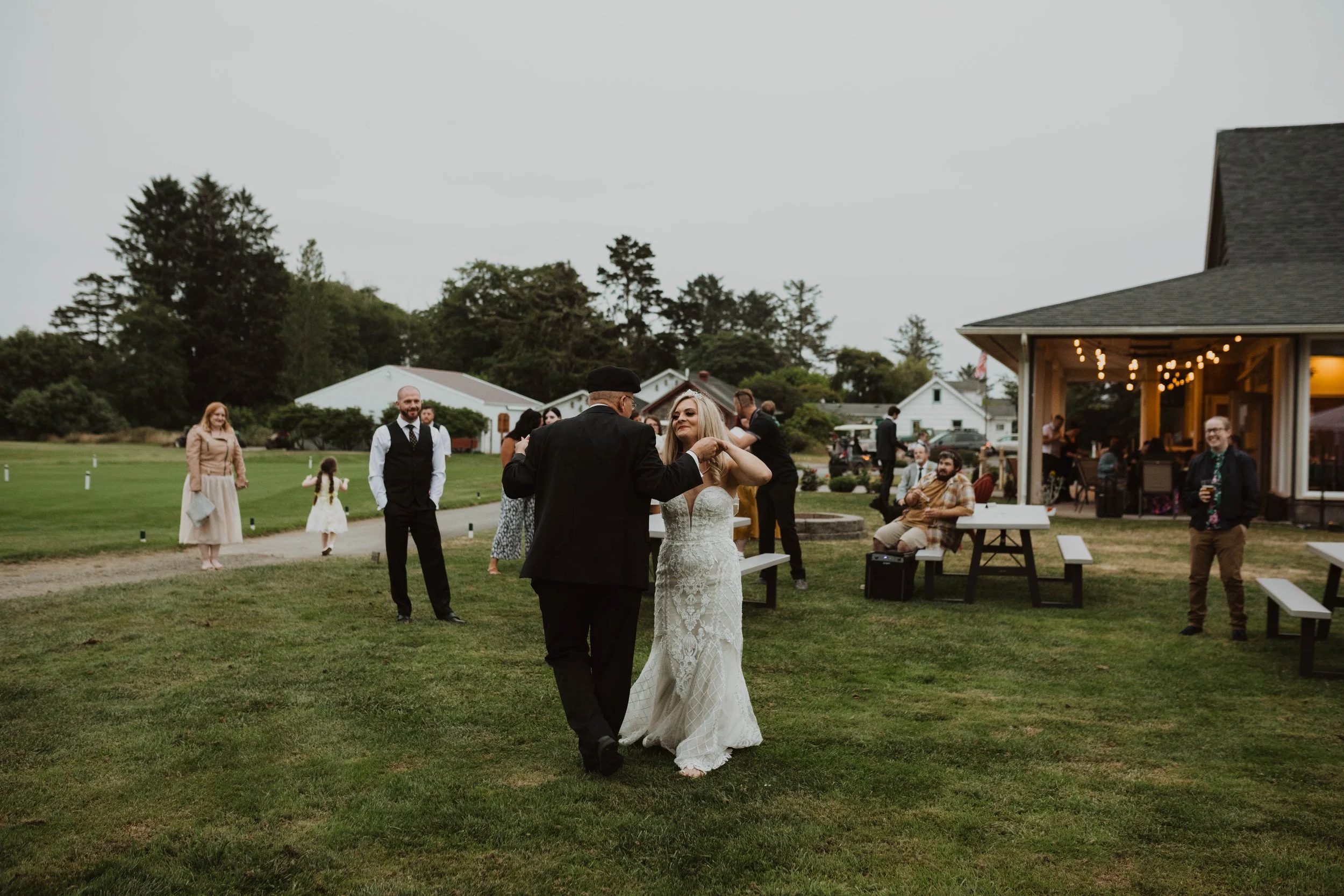 A wedding reception taking place outdoors on a grassy area. A bride in a white wedding dress is dancing with an older man in a black suit and hat. Several other guests are present, some standing and some sitting, with a building and string lights in 