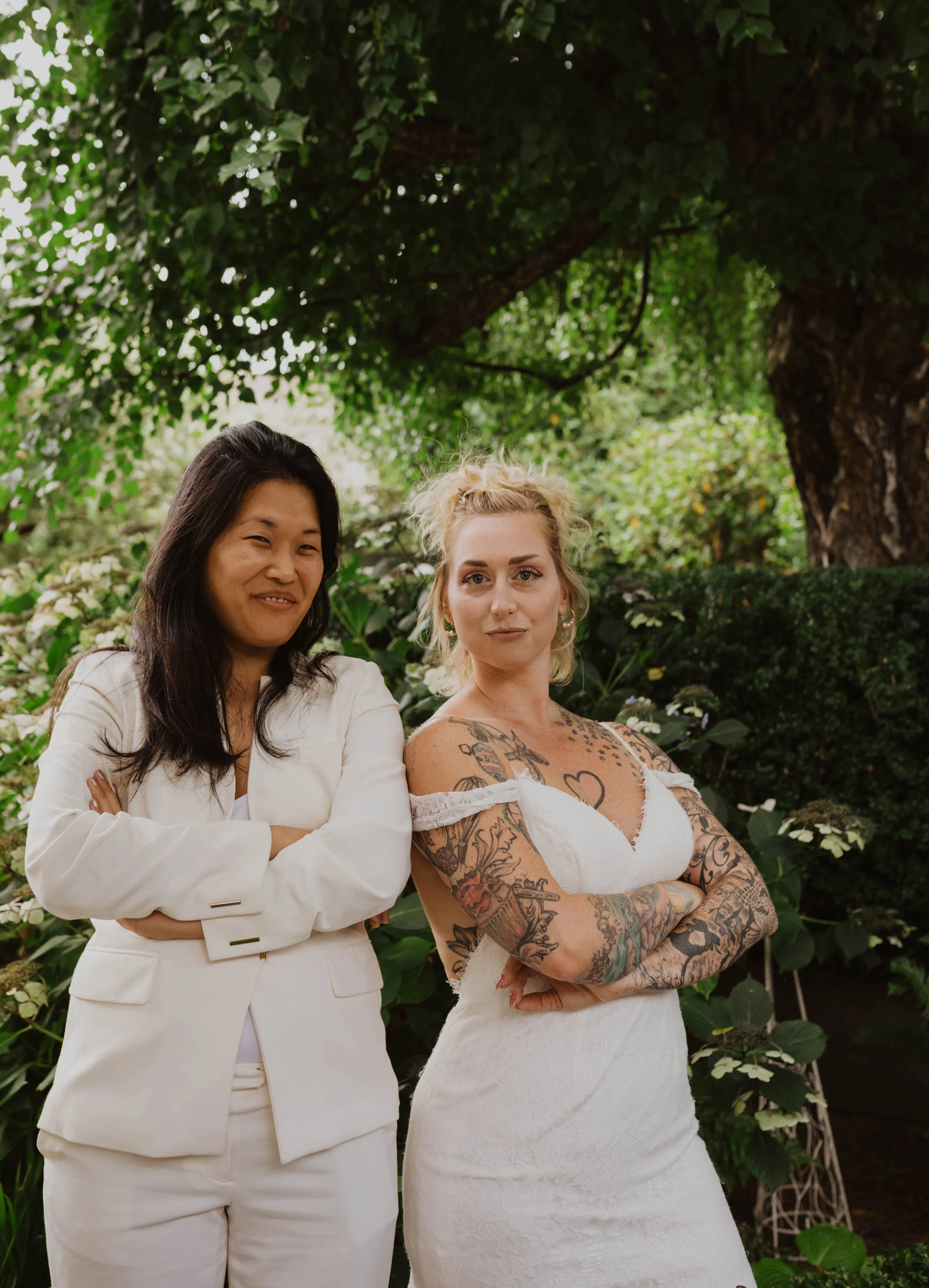 Two women standing outdoors in front of green foliage, with one woman wearing a white suit and the other a white dress, both with crossed arms and confident expressions. Seattle, WA wedding photography.