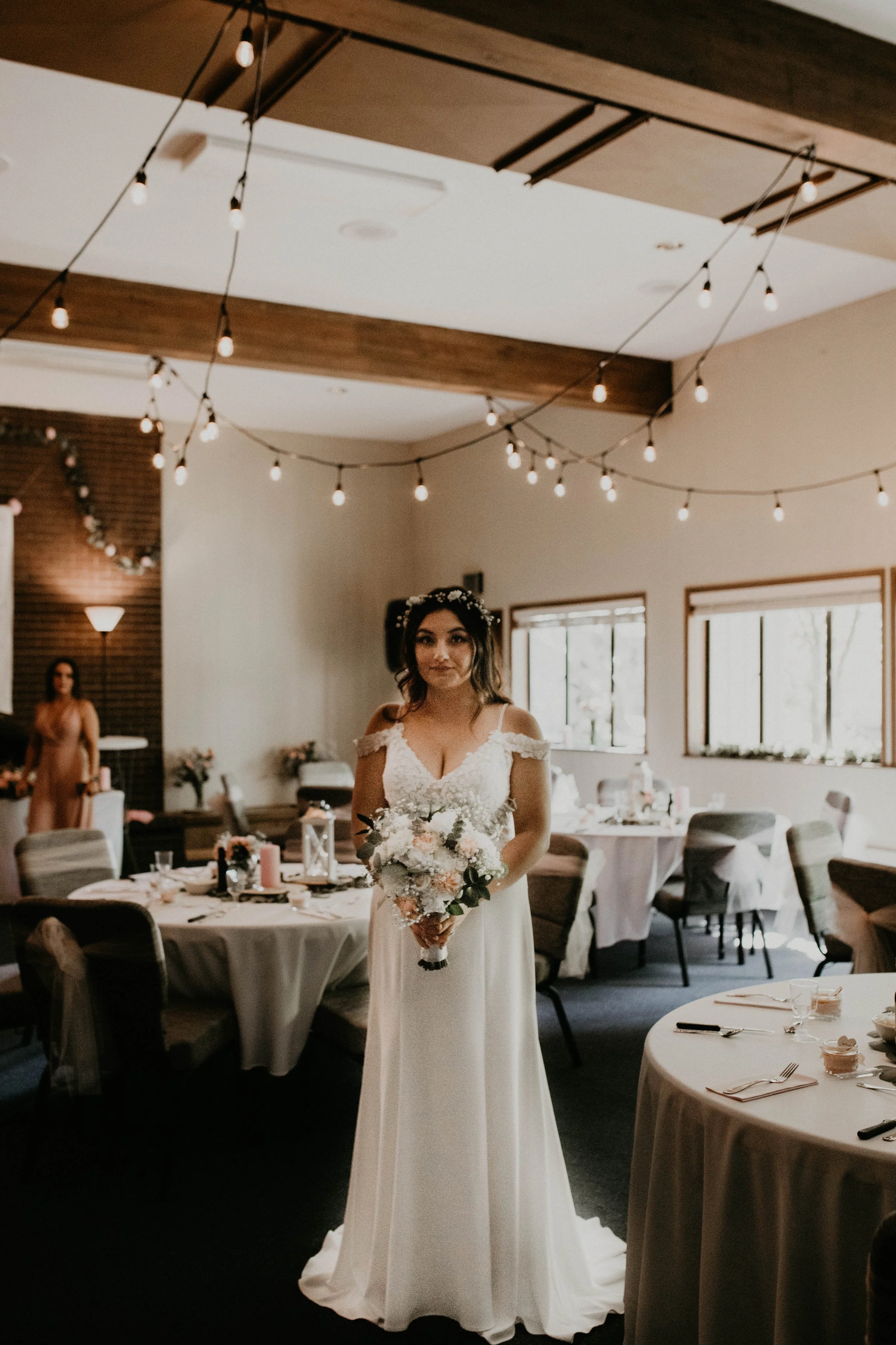 Bride in a white wedding dress holding a bouquet standing in a decorated reception hall with string lights and tables set for a celebration. Seattle, WA wedding photography.